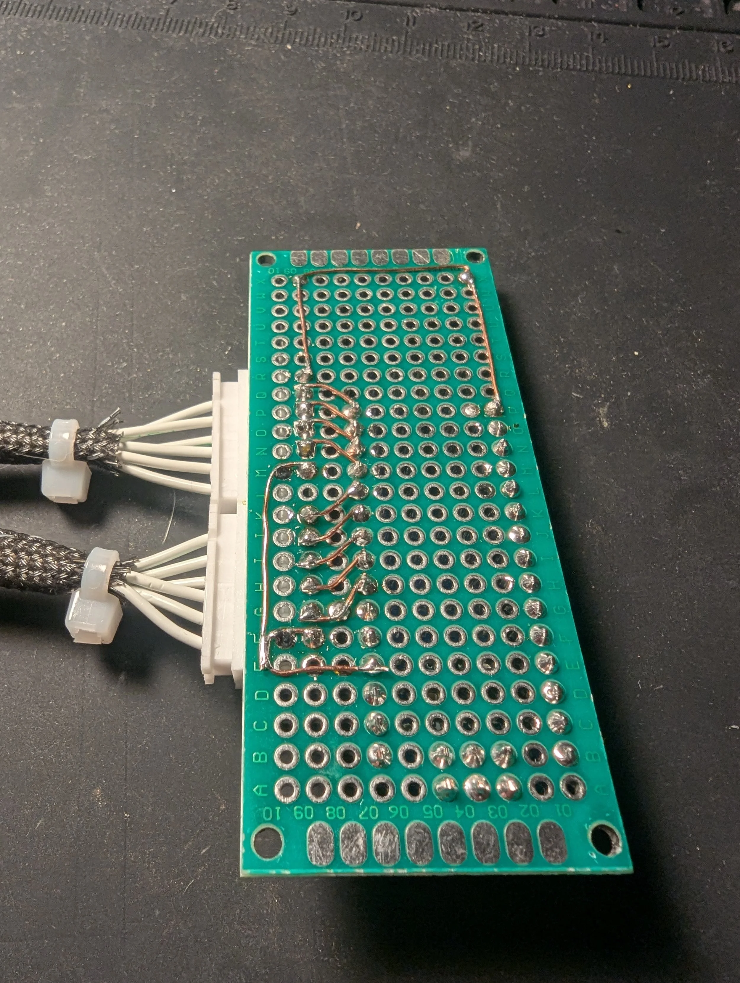 A green prototype circuit board with numerous soldered wires and holes, placed on a black work surface with a ruler in the background.