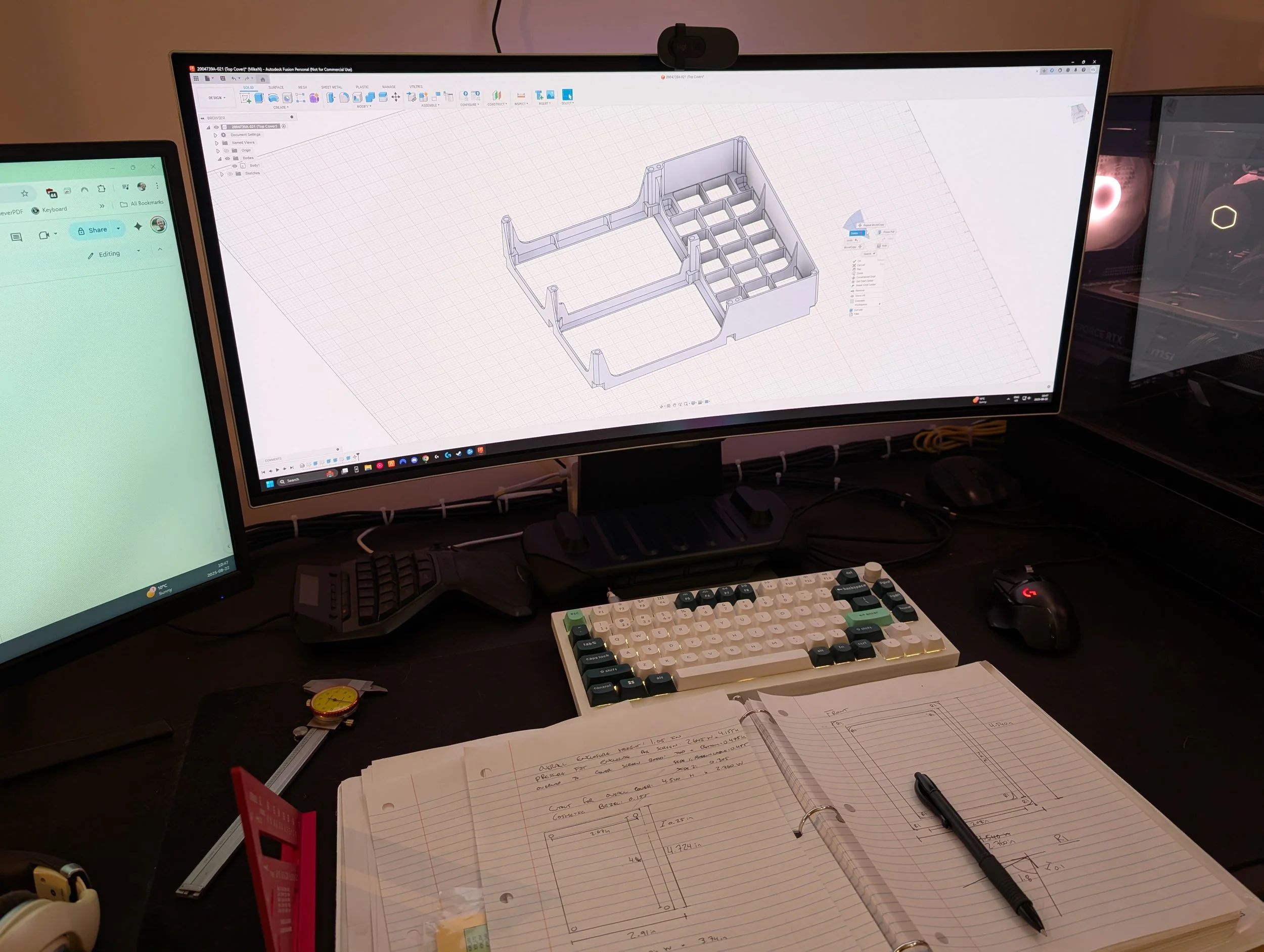 Computer desk with two monitors displaying 3D CAD design software, keyboard, mouse, notebooks with handwritten sketches and measurements, and various office supplies.