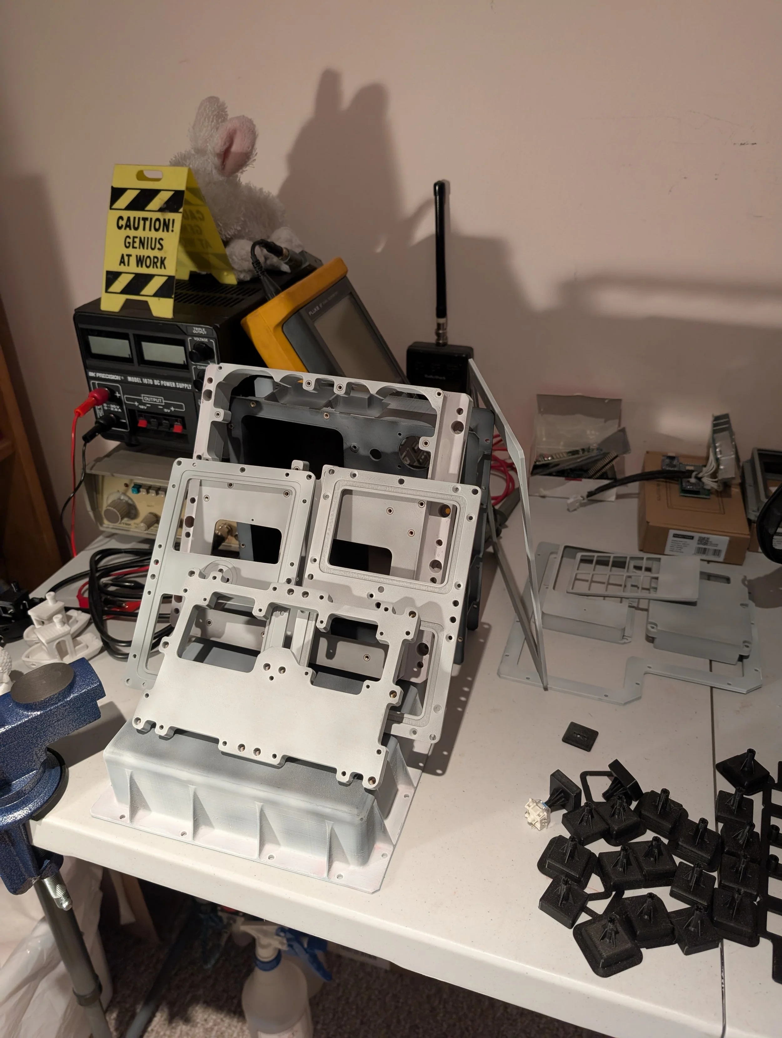 A cluttered workbench with electronic components, black keyboard keycaps, a power supply, a multimeter, and tools, set against a wall with scattered boxes and parts.