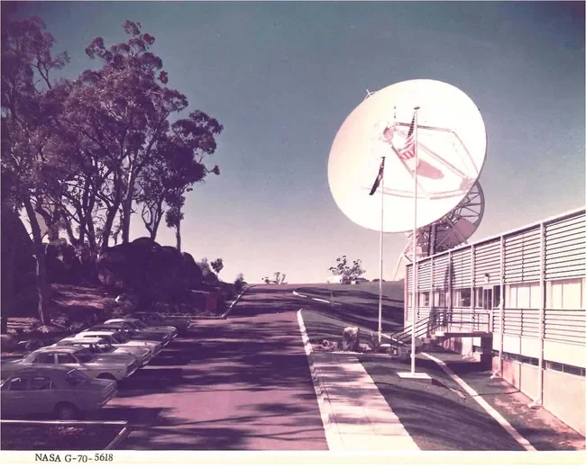 A parking lot and a large satellite dish with an American flag, trees, and a building in the background.