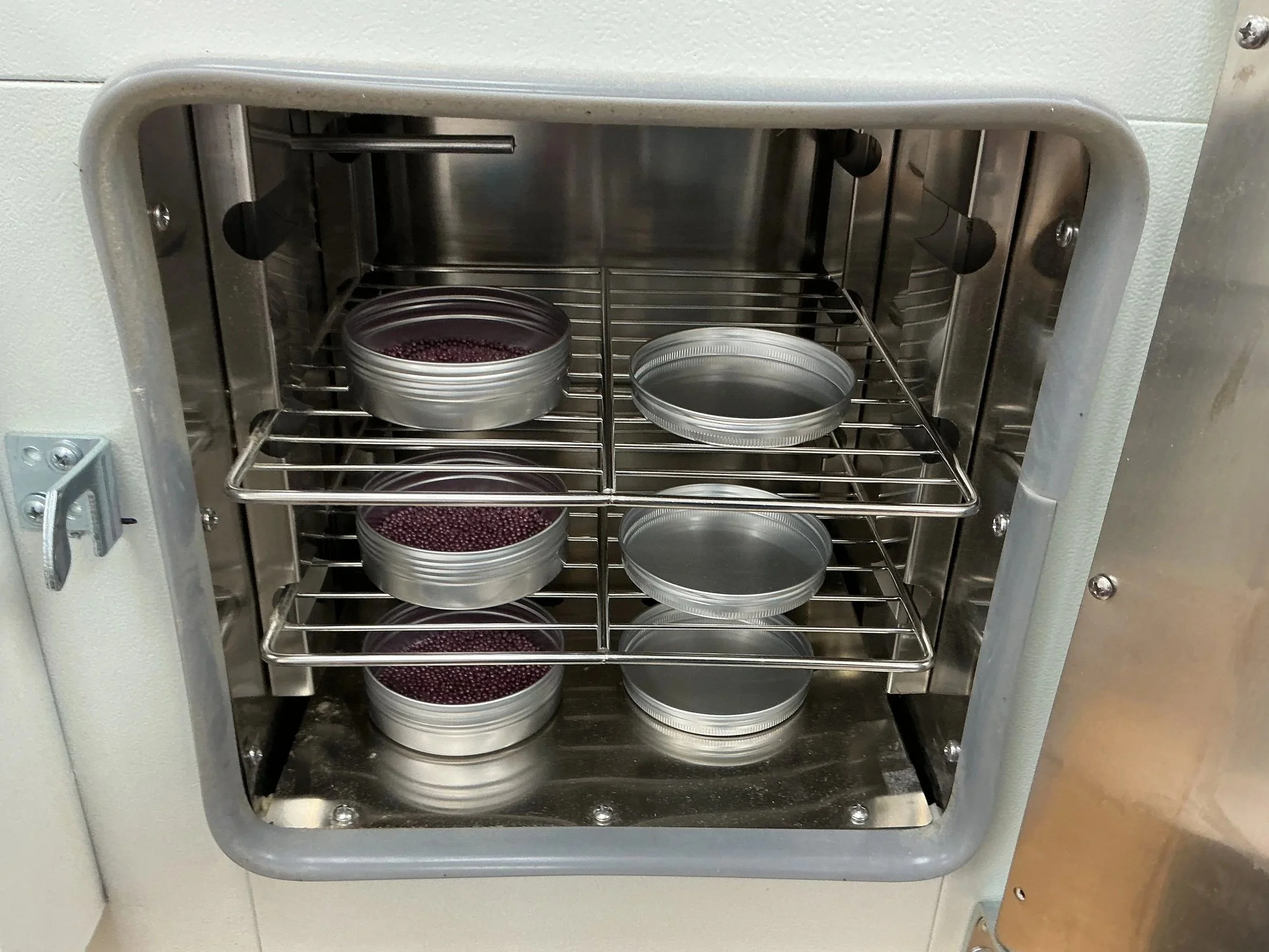 The set up of a moisture test, small metal oven with several round, silver containers on wire racks inside, some filled with canola seeds.