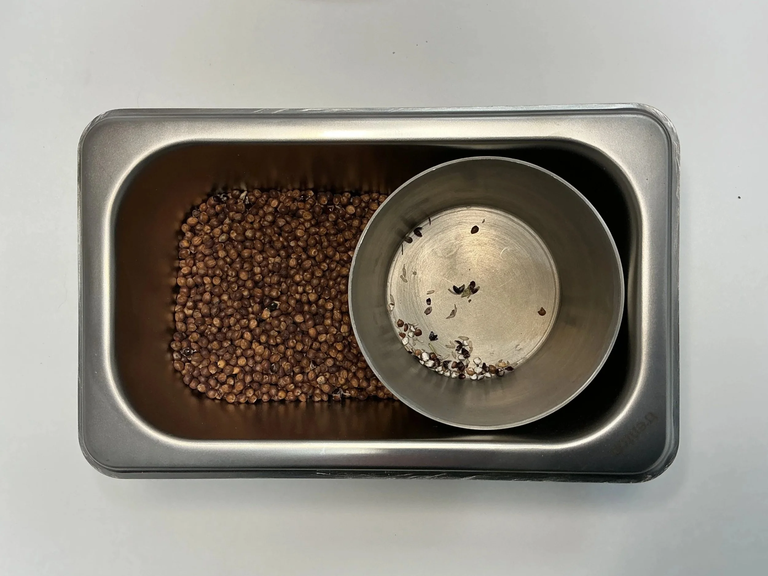 Top view of a rectangular metal container with a cylindrical metal dish inside. Pure sorghum fills the rectangular container while the impurities, inert matter, fill the dish.