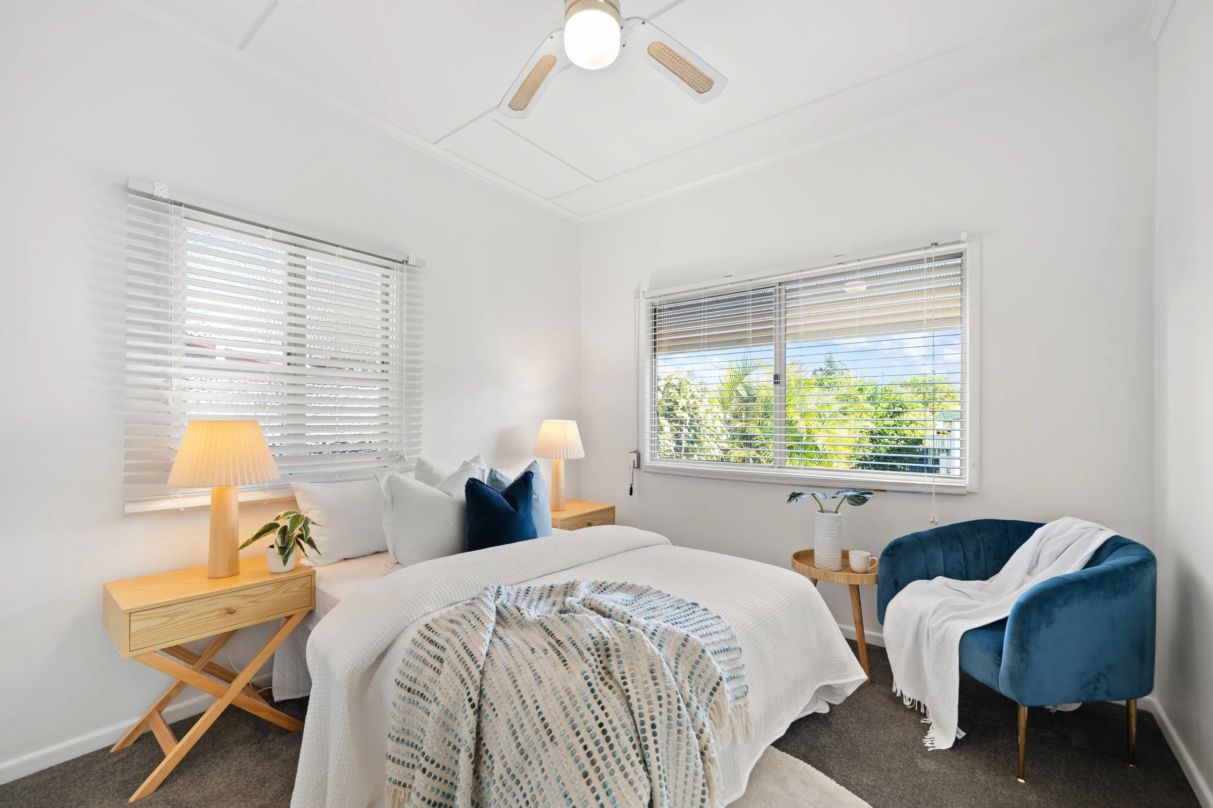 A bright bedroom with a white bed, blue and white decorative pillows, and a cream blanket. Two wooden nightstands with lamps, houseplants, and cups. Large windows with white blinds showing a green outdoor landscape. A ceiling fan with light, and grey