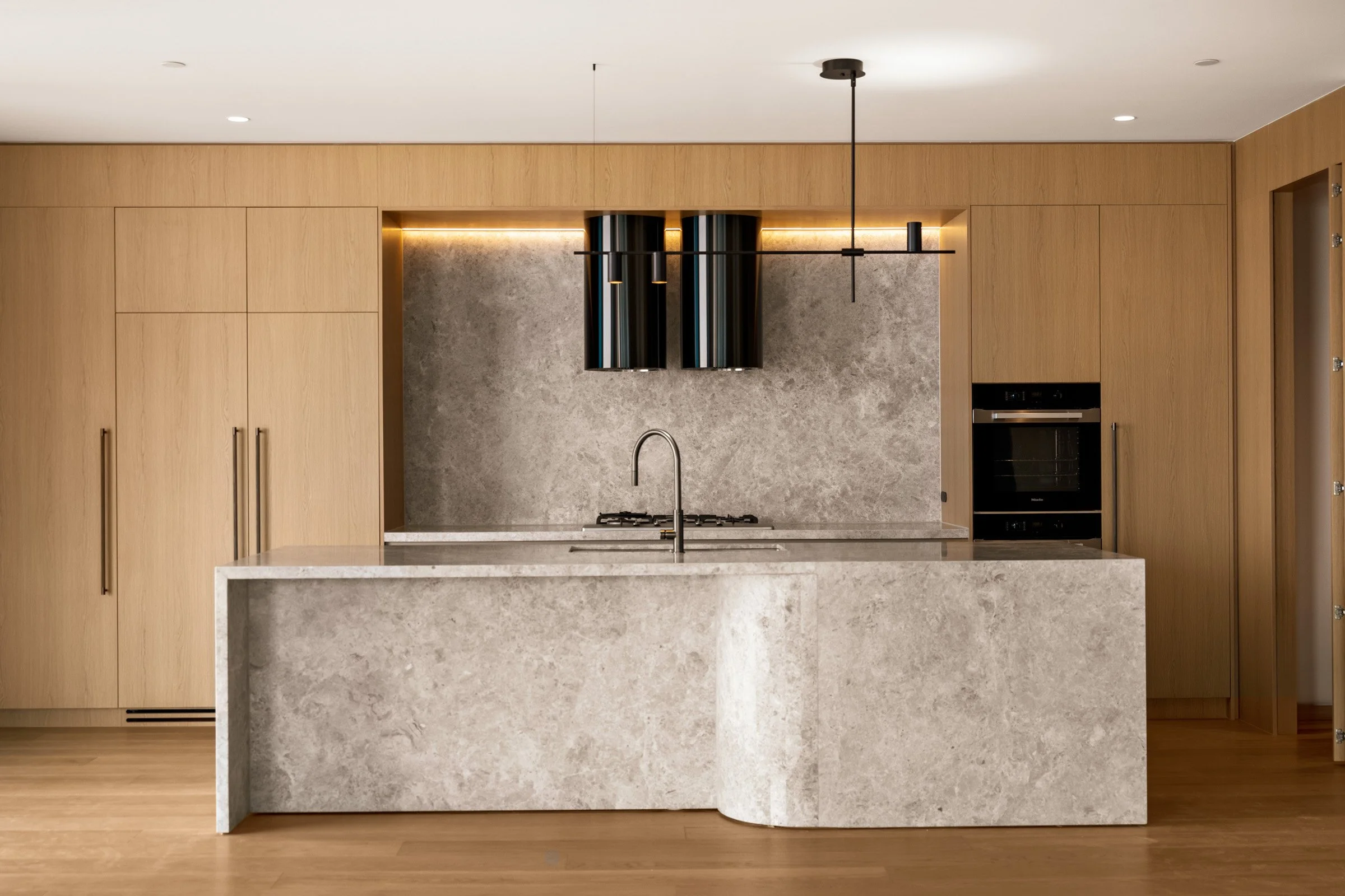 Modern kitchen with wood cabinets, a marble kitchen island, a gray stone backsplash, built-in oven, and black kitchen vent.