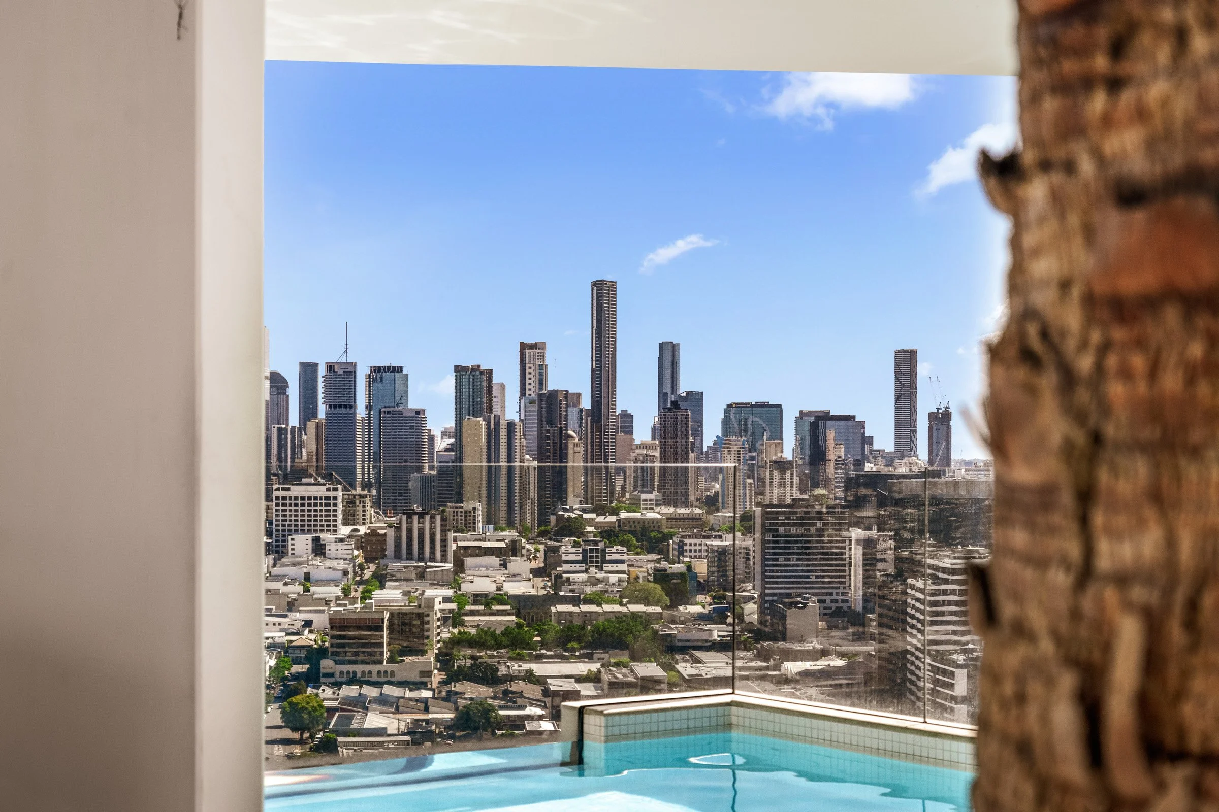 City skyline view of tall buildings from a window, with a bright blue sky and scattered clouds, partially framed by a wall and a corner of brick on the right side.
