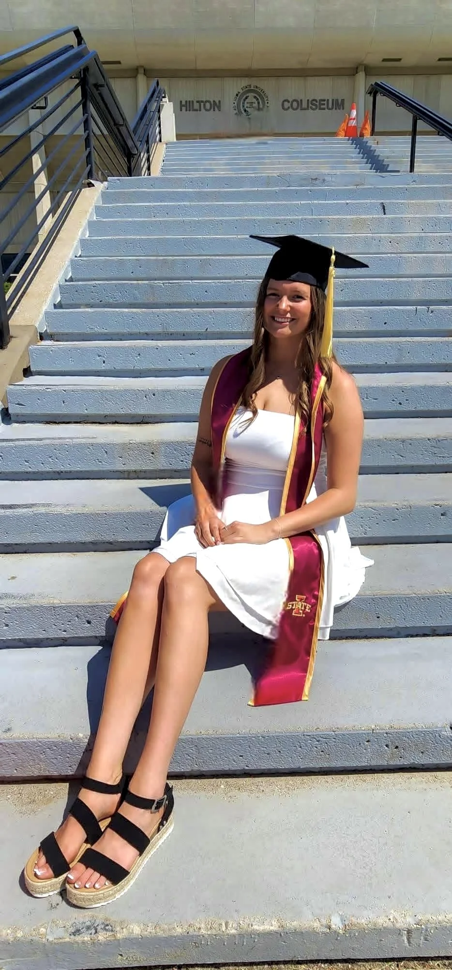 A woman wearing a cap and gown sitting on outdoor stairs, sign for Hilton Coliseum and Iowa State University in the background, sunny day.