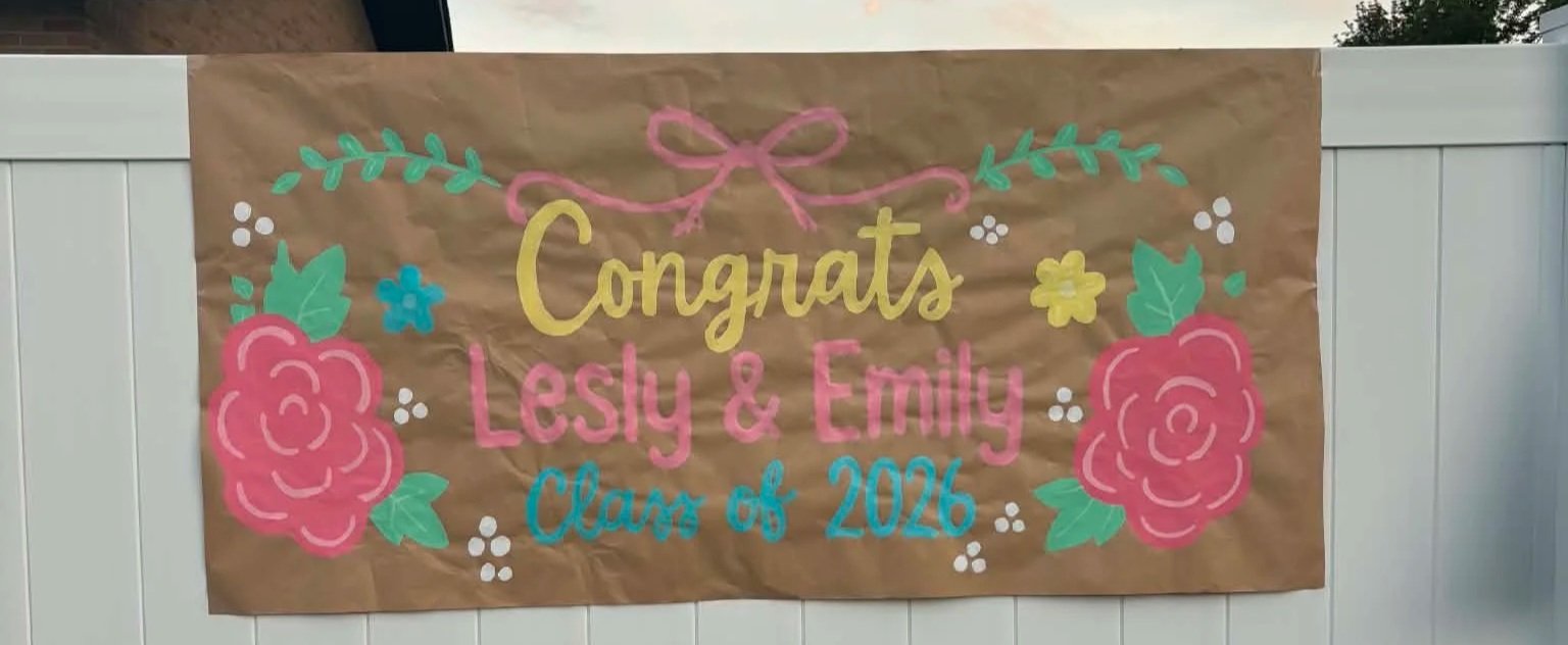 A sign on a white fence features colorful, hand-painted floral designs and text celebrating Lesly and Emily's graduation from Class of 2026.