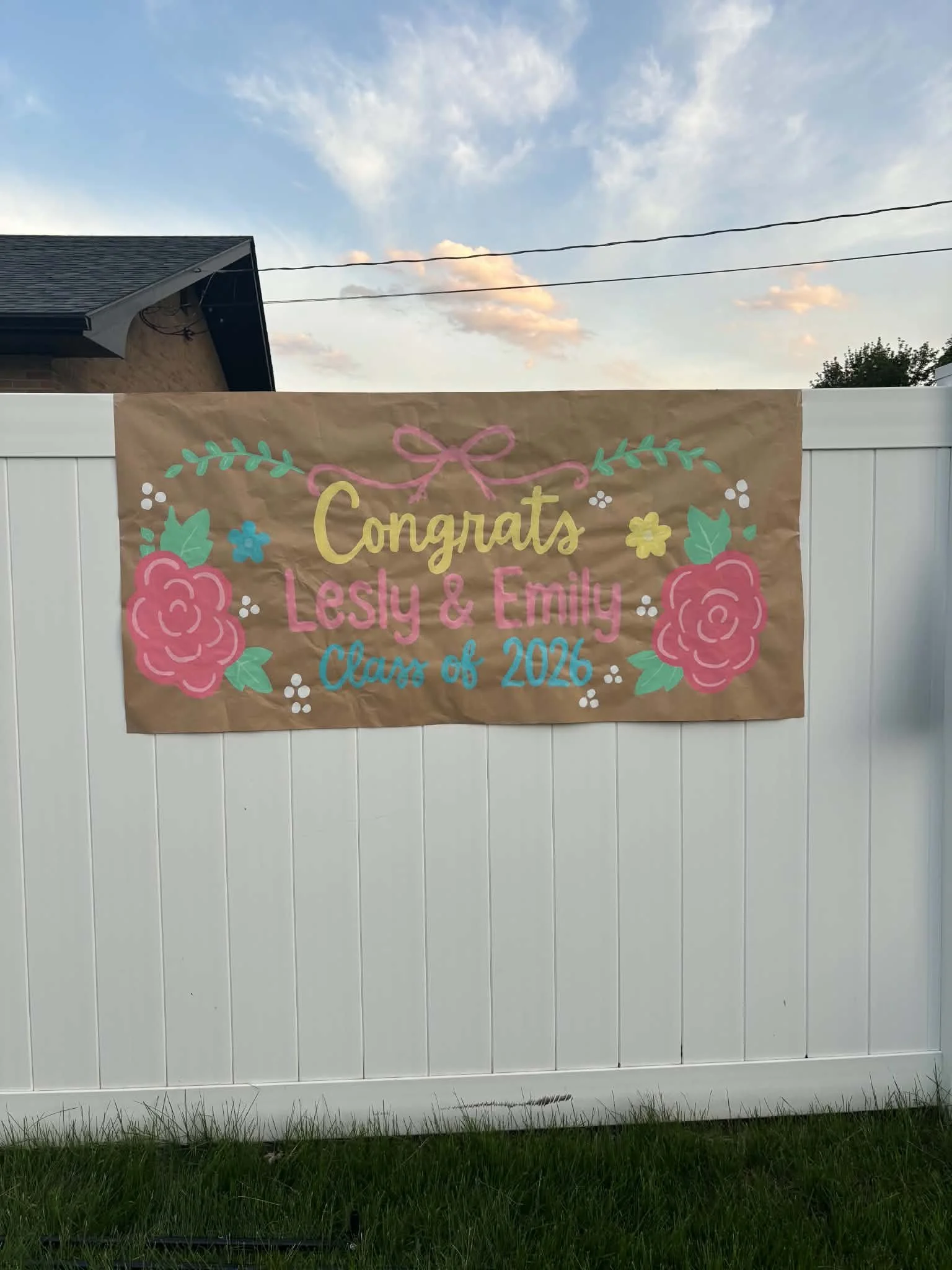 A sign on a white fence features colorful, hand-painted floral designs and text celebrating Lesly and Emily's graduation from Class of 2026.