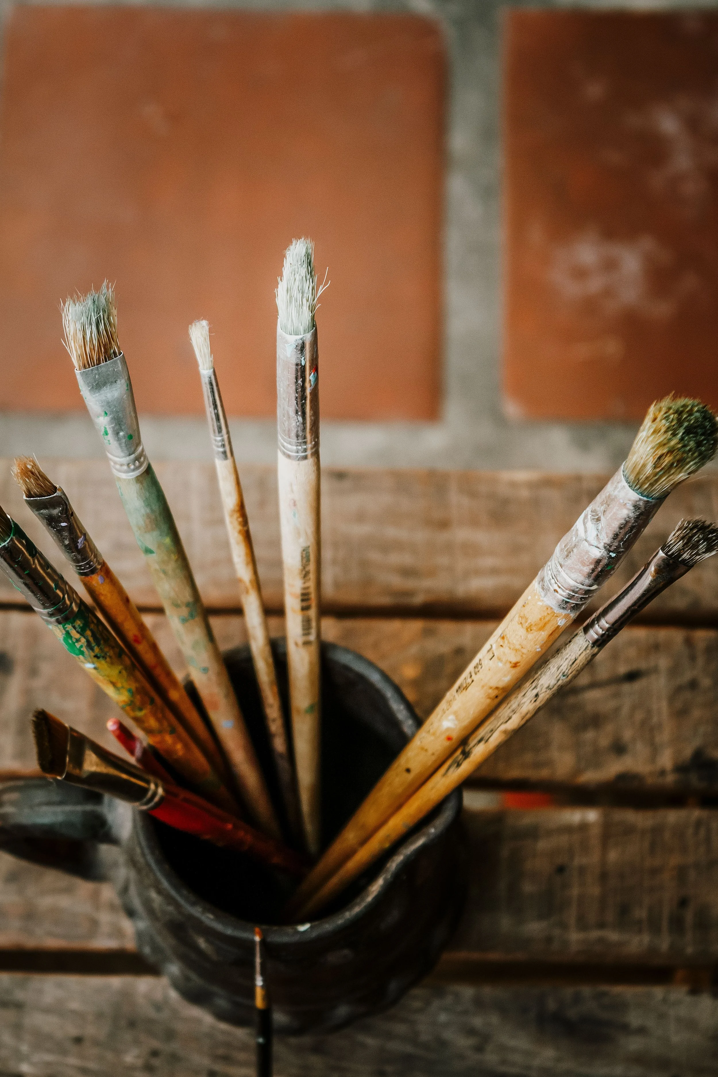 A black container holding paintbrushes of various sizes with paint residue on their handles, placed on a wooden surface.
