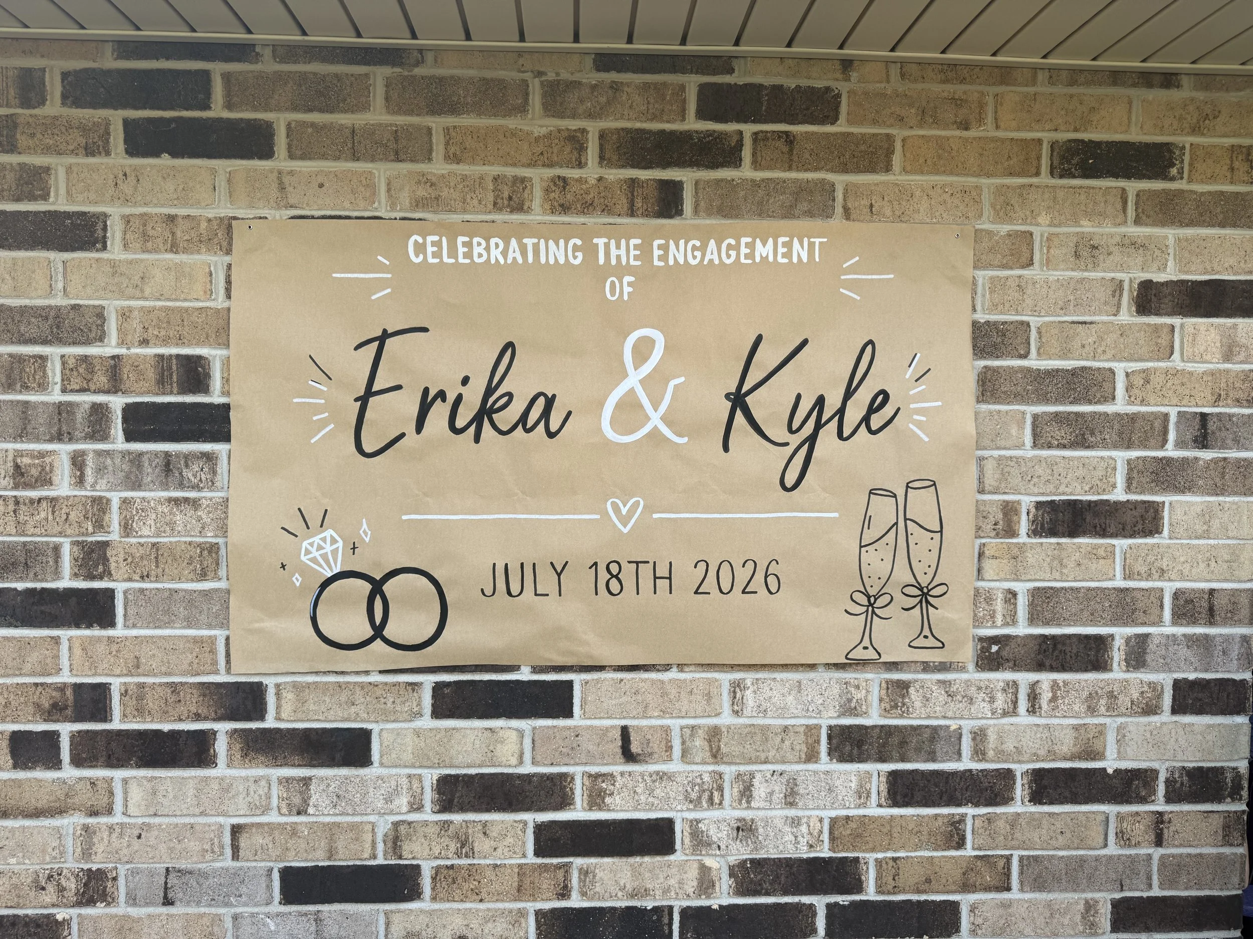 A beige poster on a brick wall celebrating the engagement of Erika and Kyle, dated July 18th, 2026, with illustrations of wedding rings, champagne glasses, and a diamond