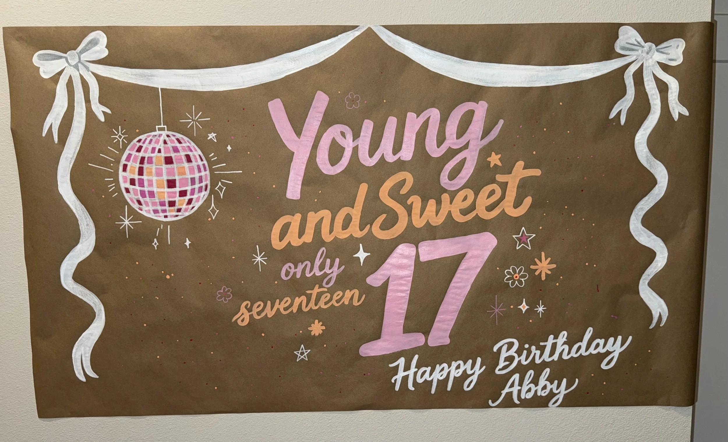 Decorative birthday sign with pink and white text, a disco ball, white ribbons, and stars on a brown background.