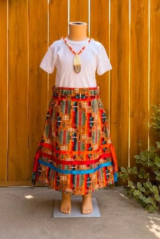A mannequin dressed in a white top, colorful beaded necklace, and a vibrant patterned skirt, displayed outdoors in front of a wooden fence.