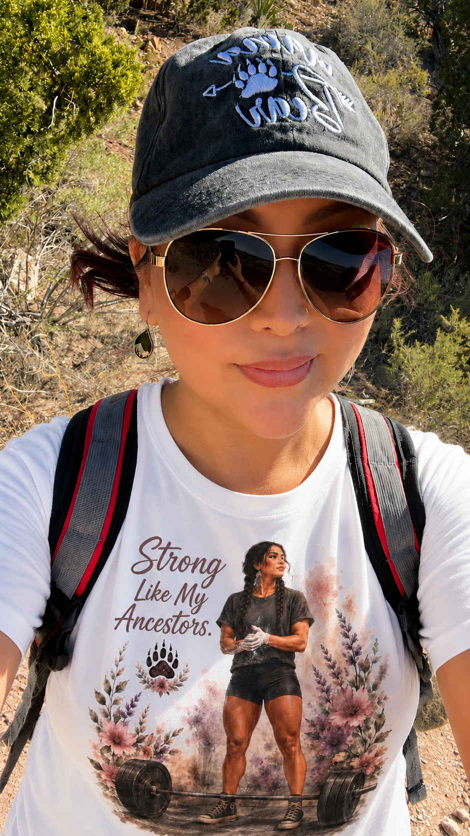 A woman outdoors wearing sunglasses, a gray baseball cap with a bear paw and mountain design, a white t-shirt with a woman lifting weights and the phrase "Strong Like My Ancestors," and a backpack. She is surrounded by greenery and desert plants.