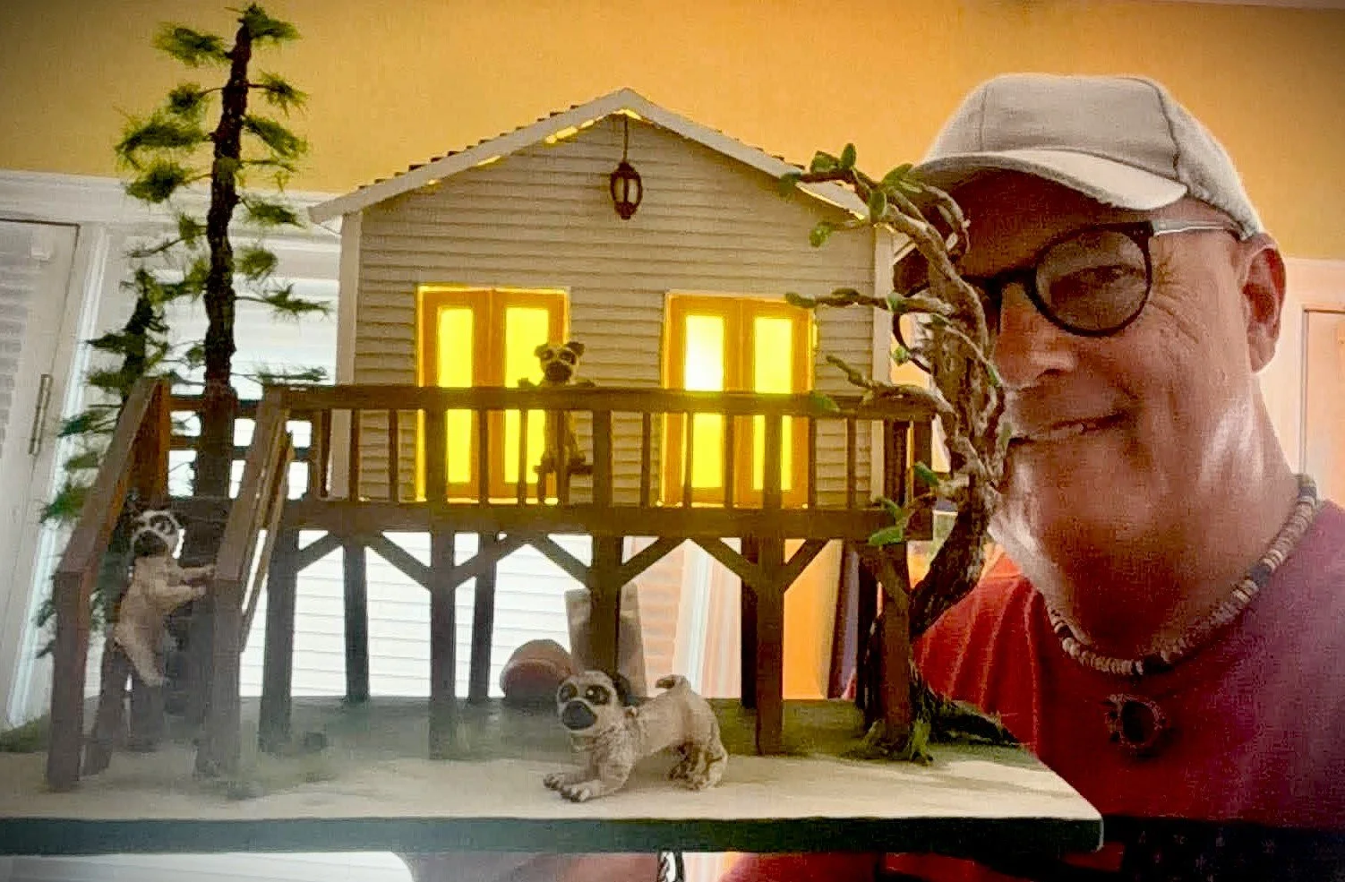 A man smiling with glasses, a cap, and a necklace next to a handcrafted miniature treehouse with three dogs, one sitting on the stairs, one standing on the porch, and one lying on the ground, all with similar markings, inside a house with a yellow-lit window background.
