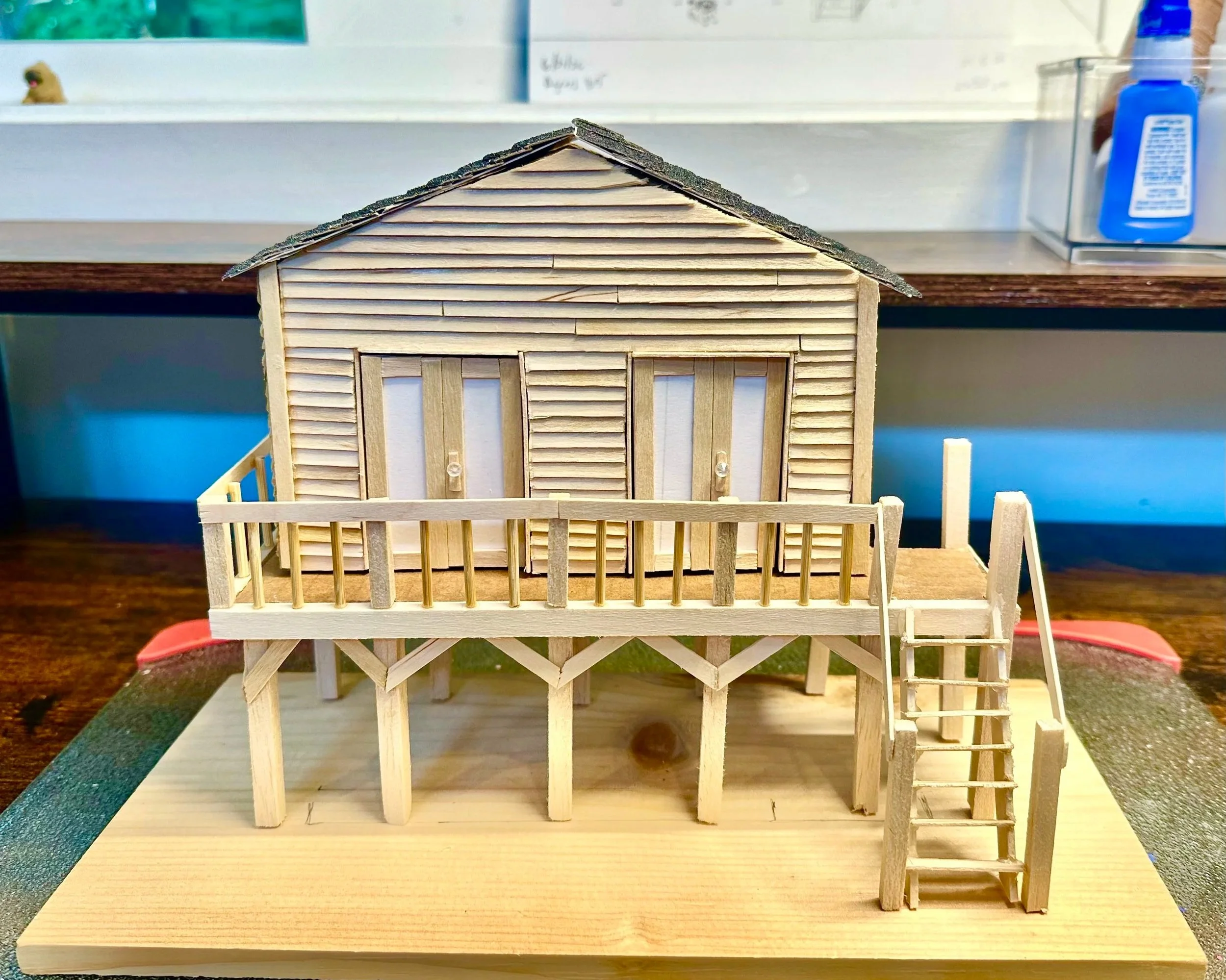 A small wooden model house with a raised foundation, wooden siding, and a gabled roof, situated on a table.
