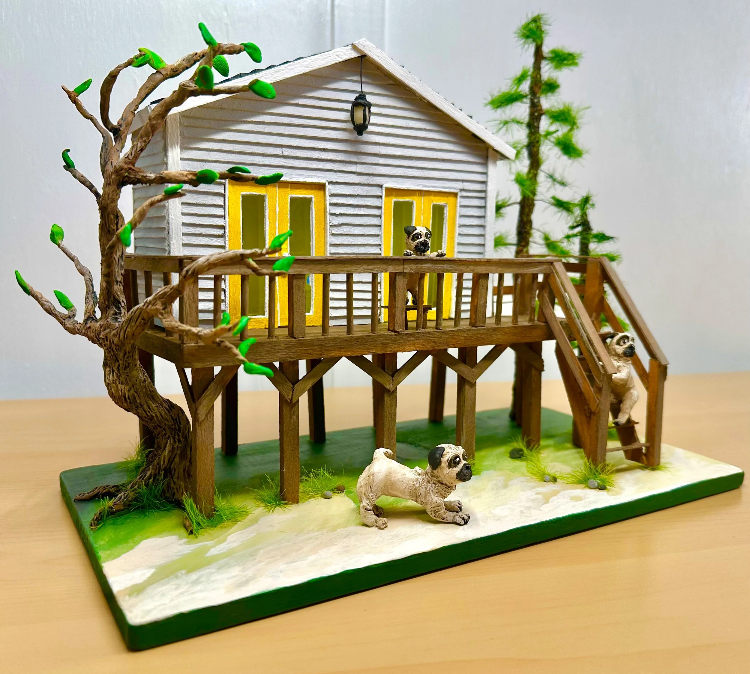 A detailed miniature model of a house on a green base, with a tree and two dogs, one standing outside near the house and one peeking from behind the house.