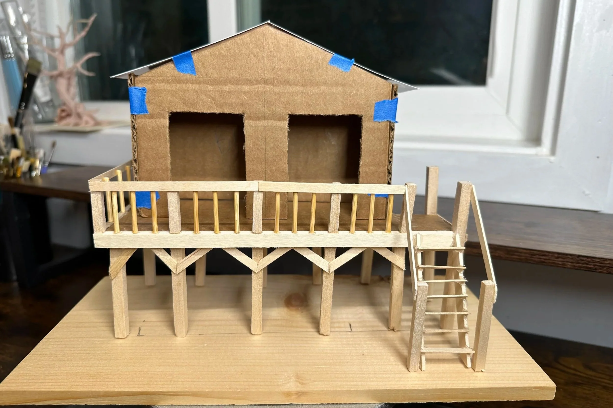 A scale model of a house on a wooden surface, made of cardboard and wood, with a small balcony and stairs, located indoors near a window.