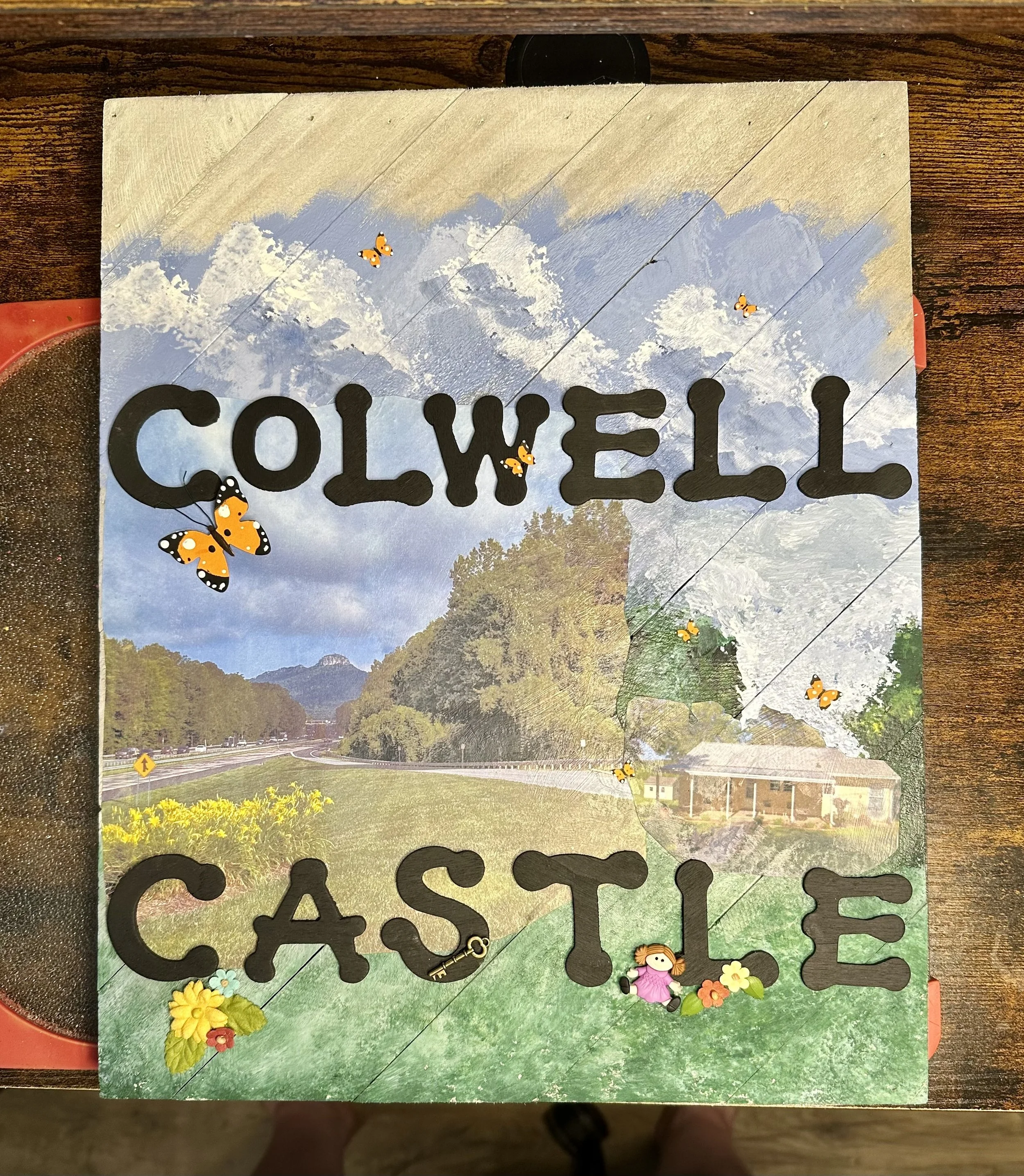 A decorative sign with the words 'Welcome Castle' in large black letters, adorned with butterfly stickers, on a outdoor background with trees and a road.