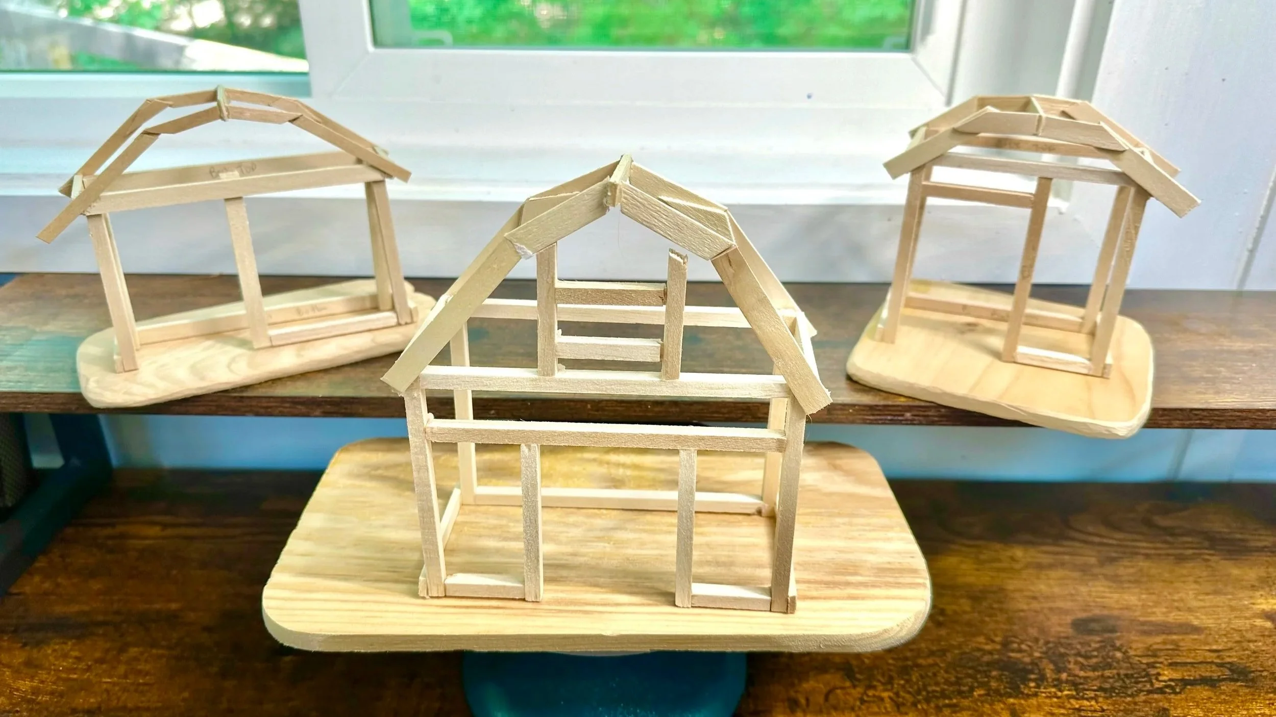Three small wooden house models in progress, set on individual wooden bases, with various frame structures displayed near a window.