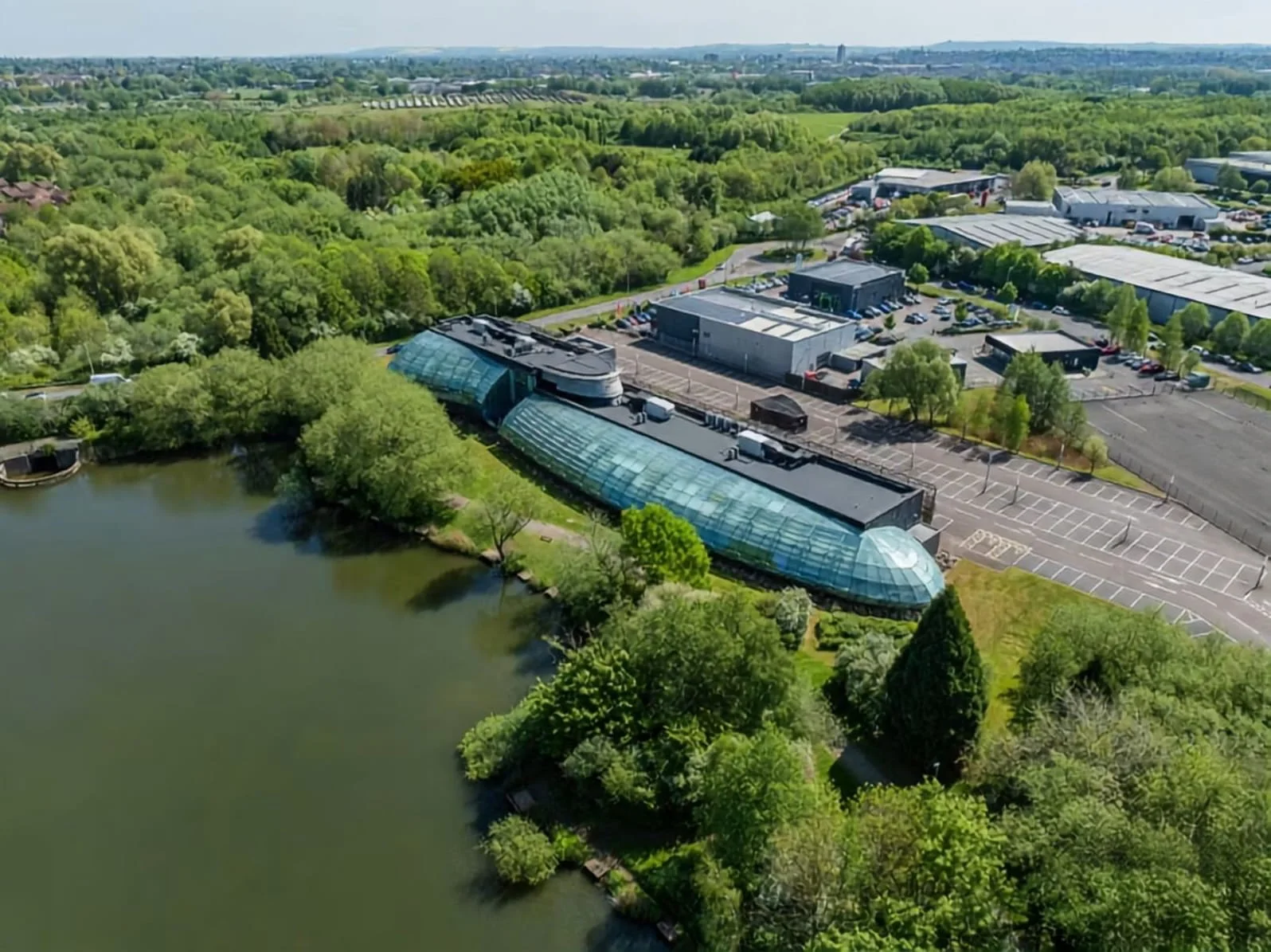 Aerial view of a park with a lake, surrounded by lush green trees, and a modern building with a glass roof near the water. There are parking lots and commercial buildings in the background.