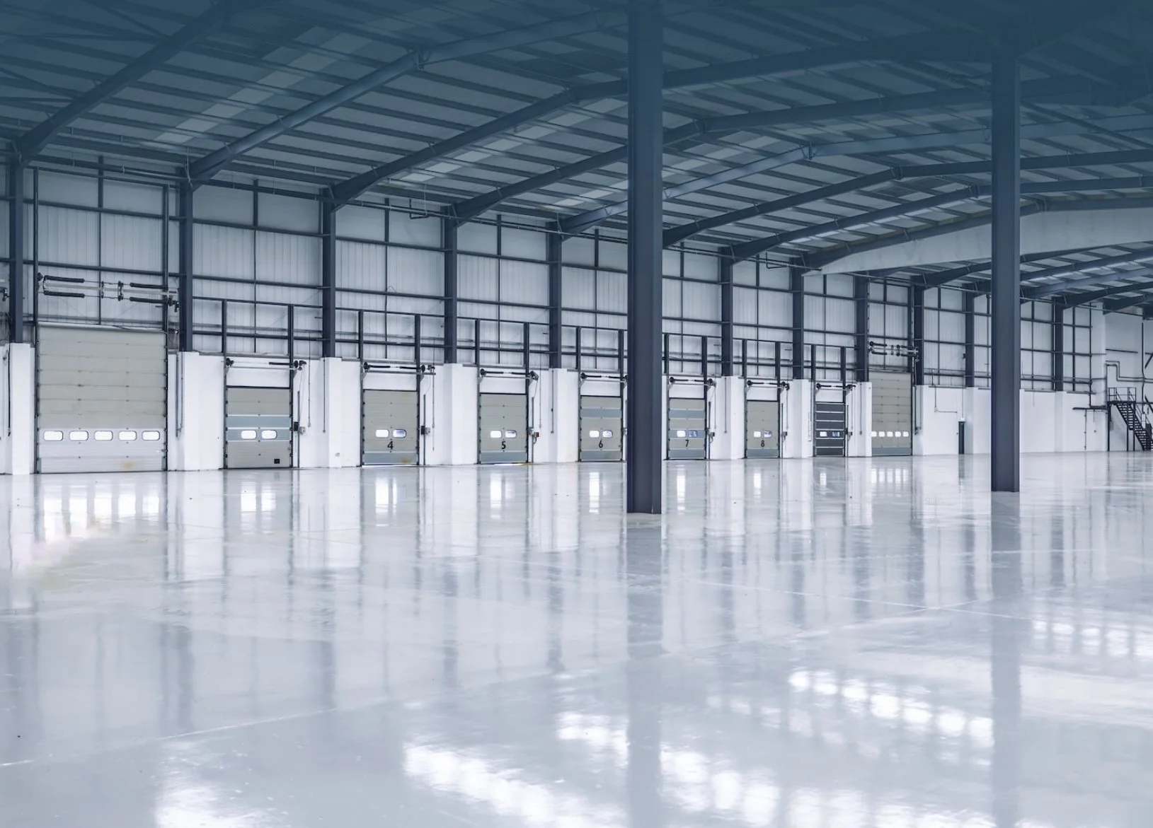 Empty warehouse with high ceilings, large garage doors numbered 4 to 8, and a polished concrete floor reflecting light.