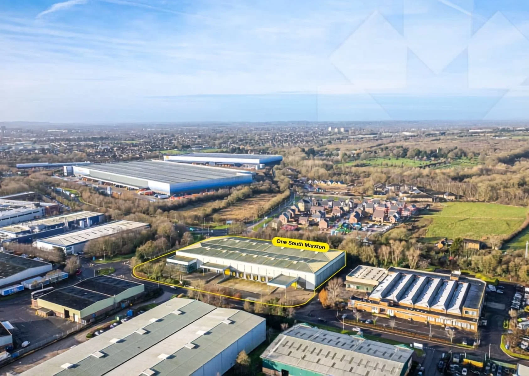 Aerial view of an industrial area near residential neighborhoods, with a large warehouse and commercial buildings, and a labeled lot called One South Marston, with trees and fields extending into the distance under a blue sky.