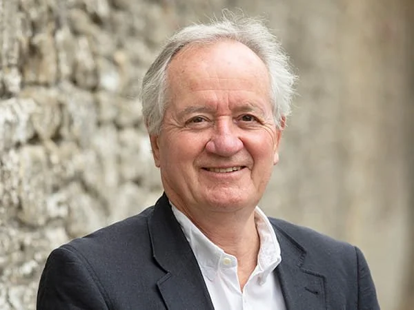 A smiling man with gray hair, wearing a dark blazer and white shirt, standing outdoors in front of a stone wall. Philip Loveday, Loveday Consultant.