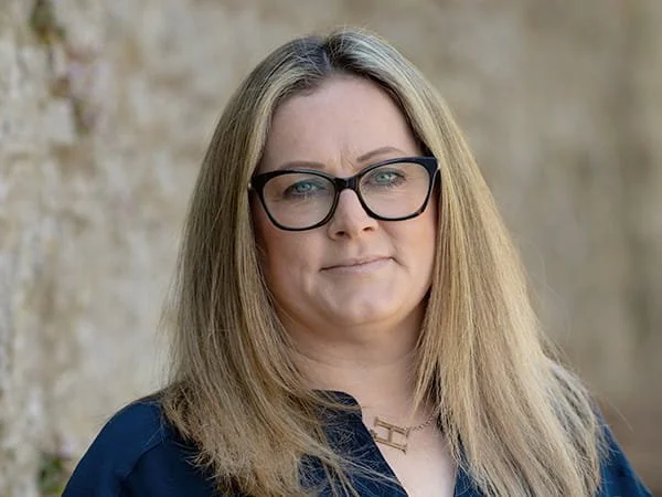 A woman with blonde hair, glasses, and a blue shirt looking at the camera outdoors. Hannah Webster, Loveday Administrator.