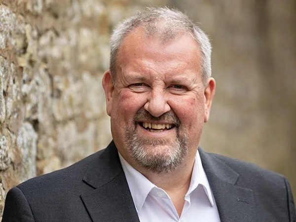 A smiling man with short gray hair, beard, and mustache, wearing a dark suit jacket and white dress shirt, standing outdoors against a stone wall. Huw Thomas, Loveday Consultant.