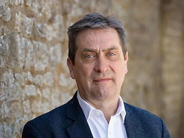 A man with short dark hair and blue eyes wearing a white shirt and dark blazer, standing outdoors with a stone wall background. Peter Cole, Loveday Partner.