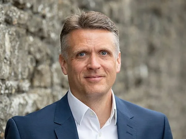 A man in a blue suit and white shirt standing outdoors in front of a stone wall. Alastair Andrews, Loveday Partner.