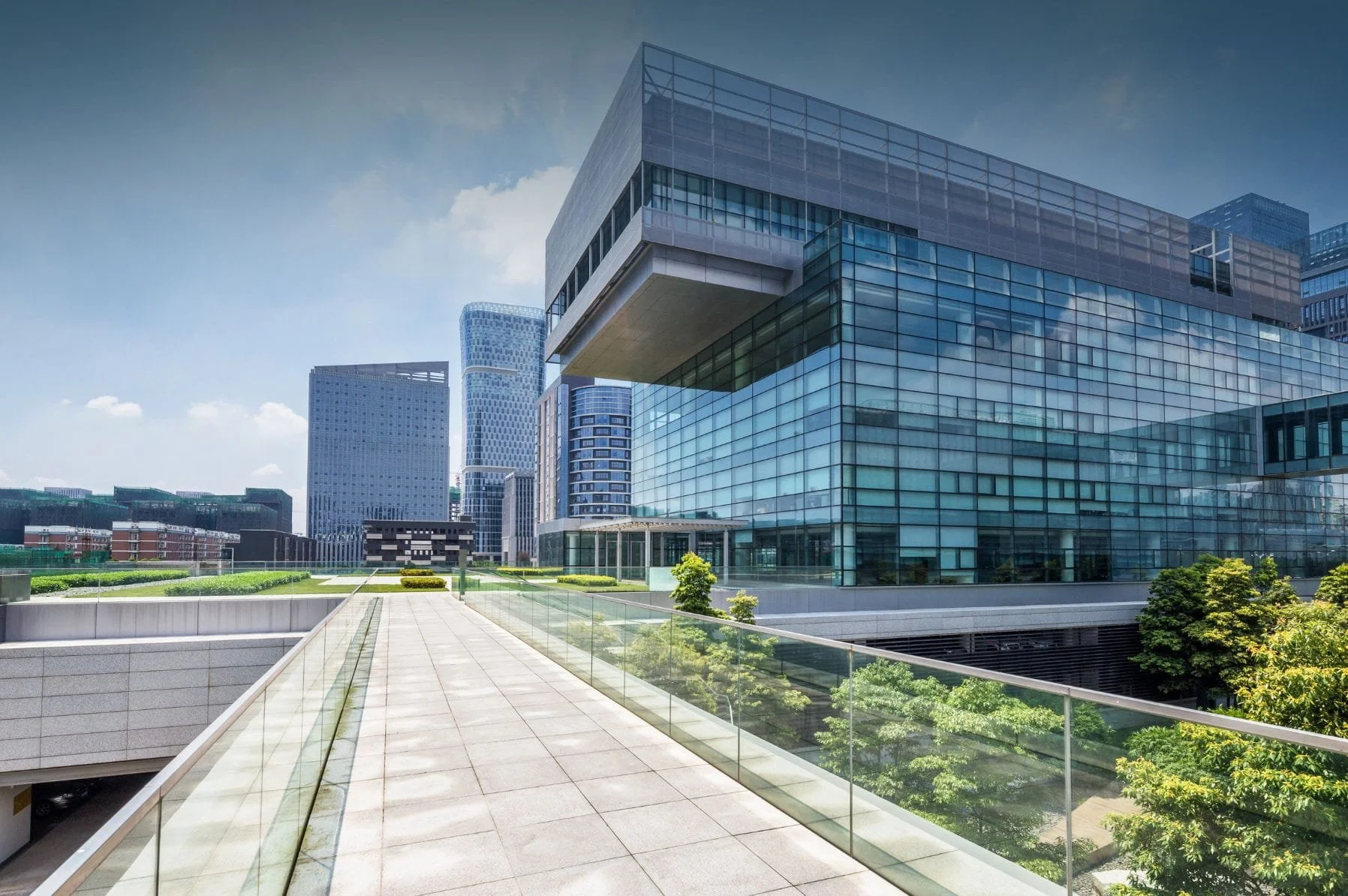Modern glass office building with a walkway and green landscaping in an urban cityscape.