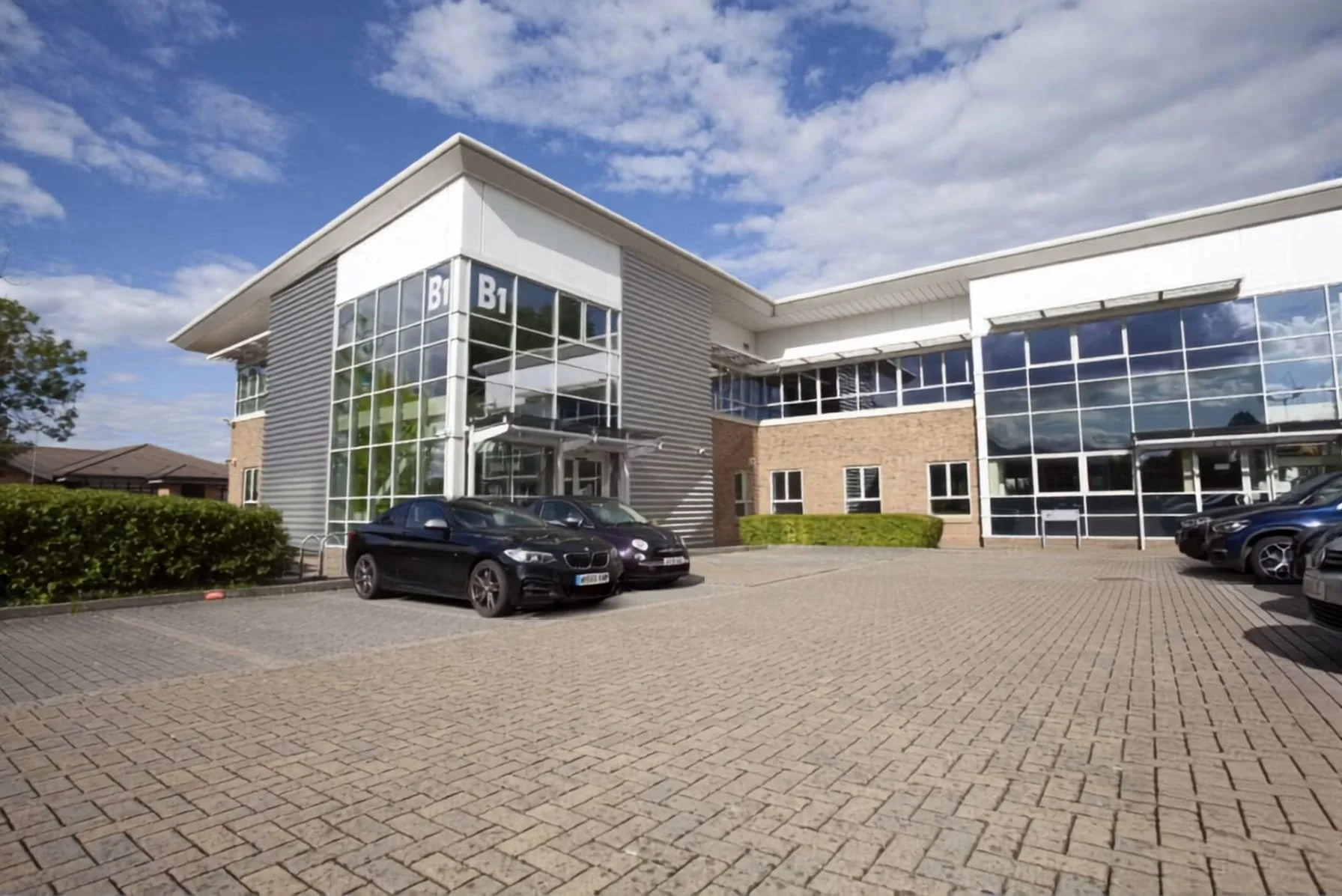 Modern office building with large glass windows, a parking lot with several cars, and a partly cloudy sky.