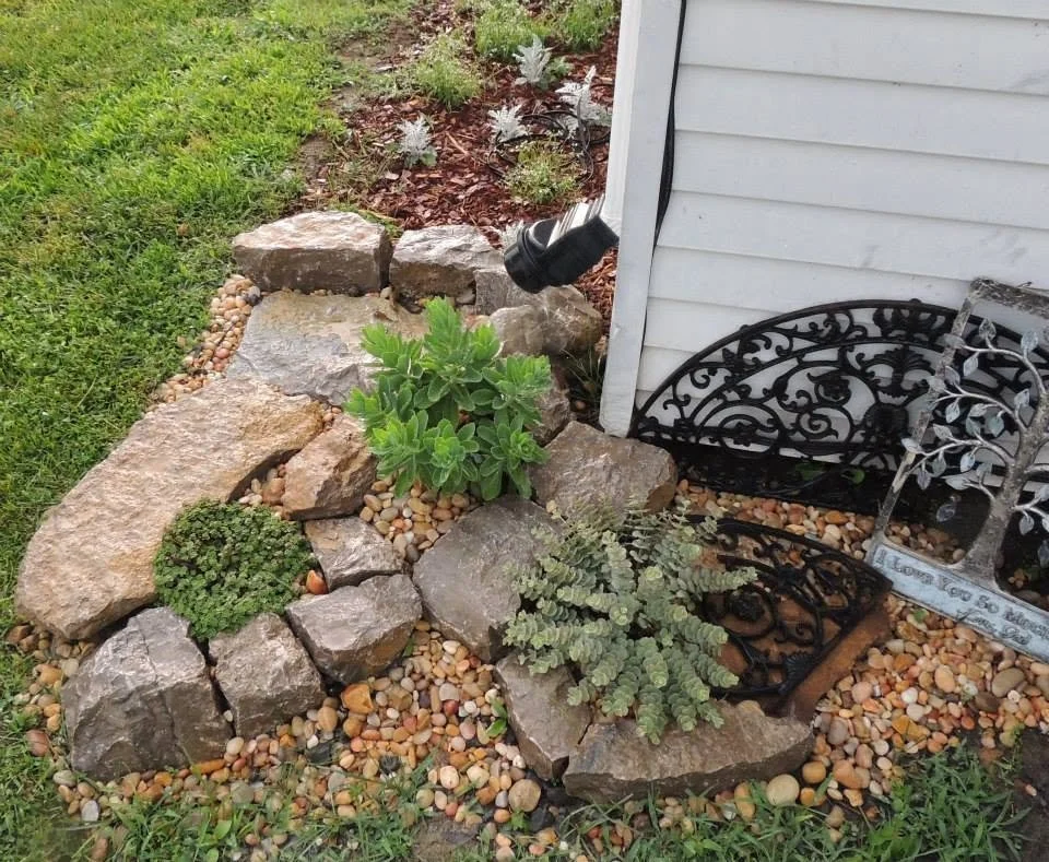 A garden bed with large rocks, green plants, and small pebbles, situated next to a house with white siding. There are decorative black metal pieces and outdoor lighting fixtures nearby.