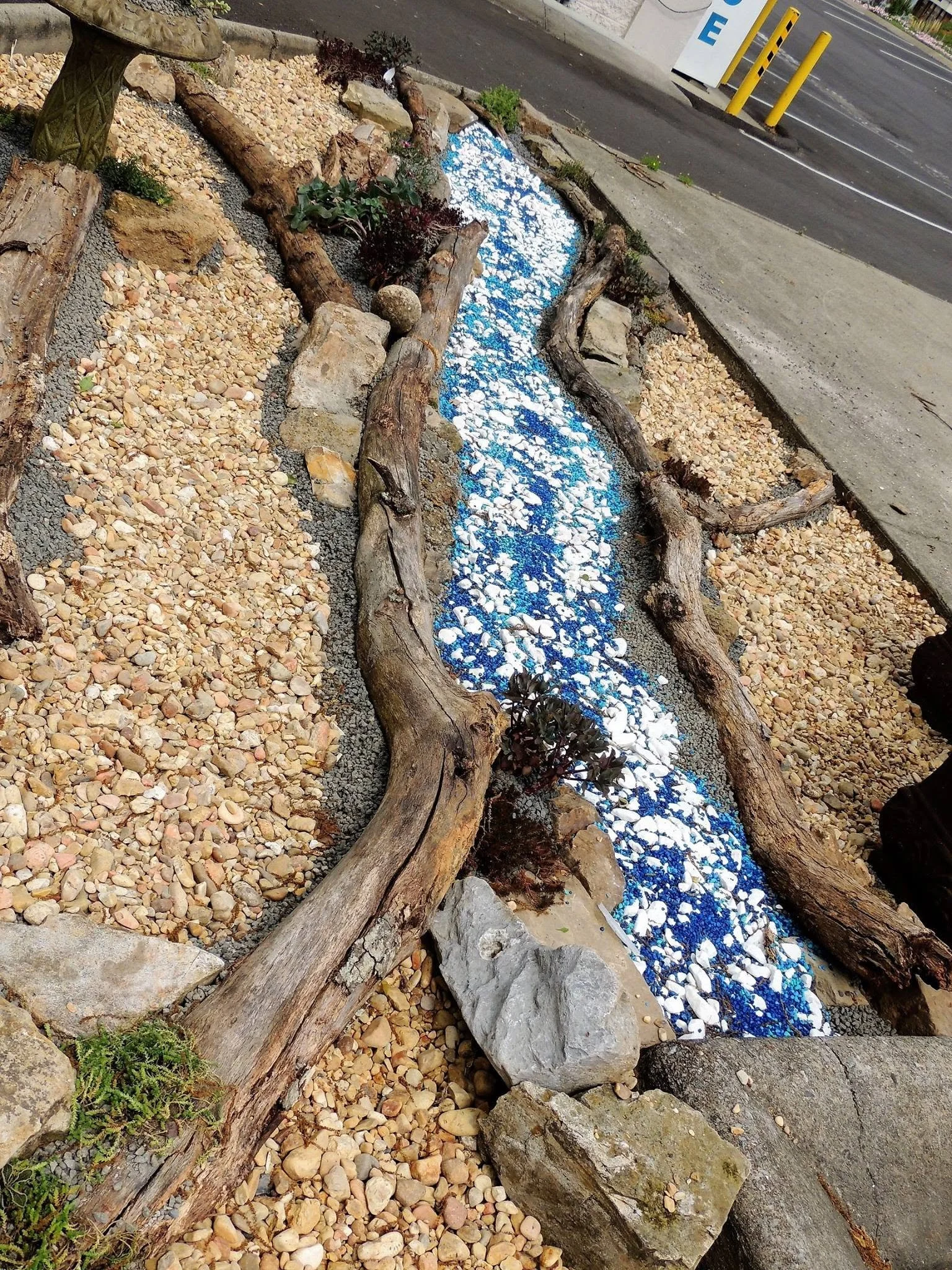 Small landscaped area with a miniature waterfall feature, surrounded by rocks and wood logs, with gravel and small plants around it, adjacent to a parking lot and sidewalk.