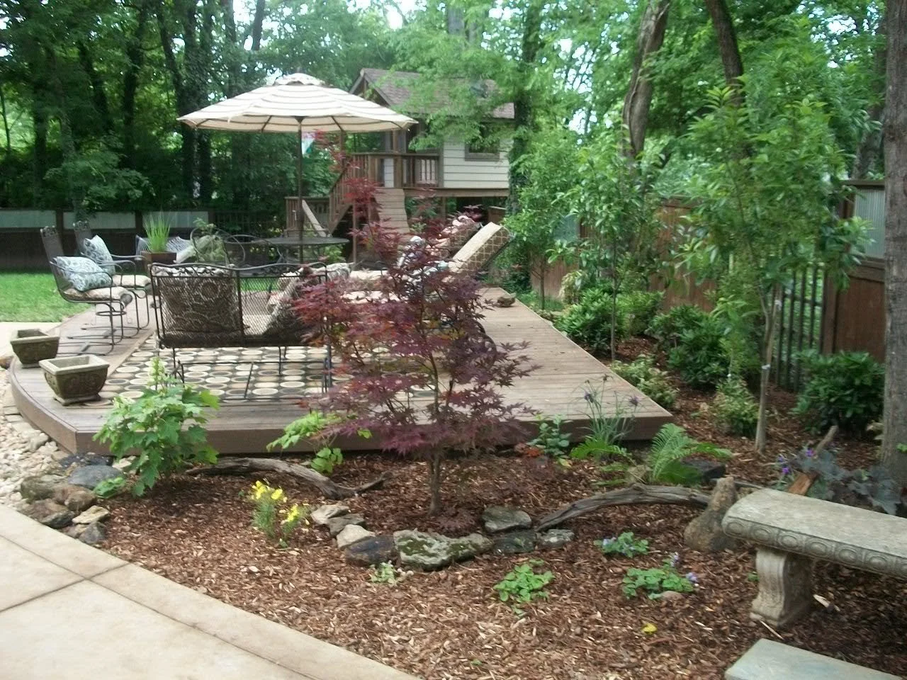 A backyard patio with outdoor furniture and a large umbrella, surrounded by trees and landscaped garden with plants and flowers.
