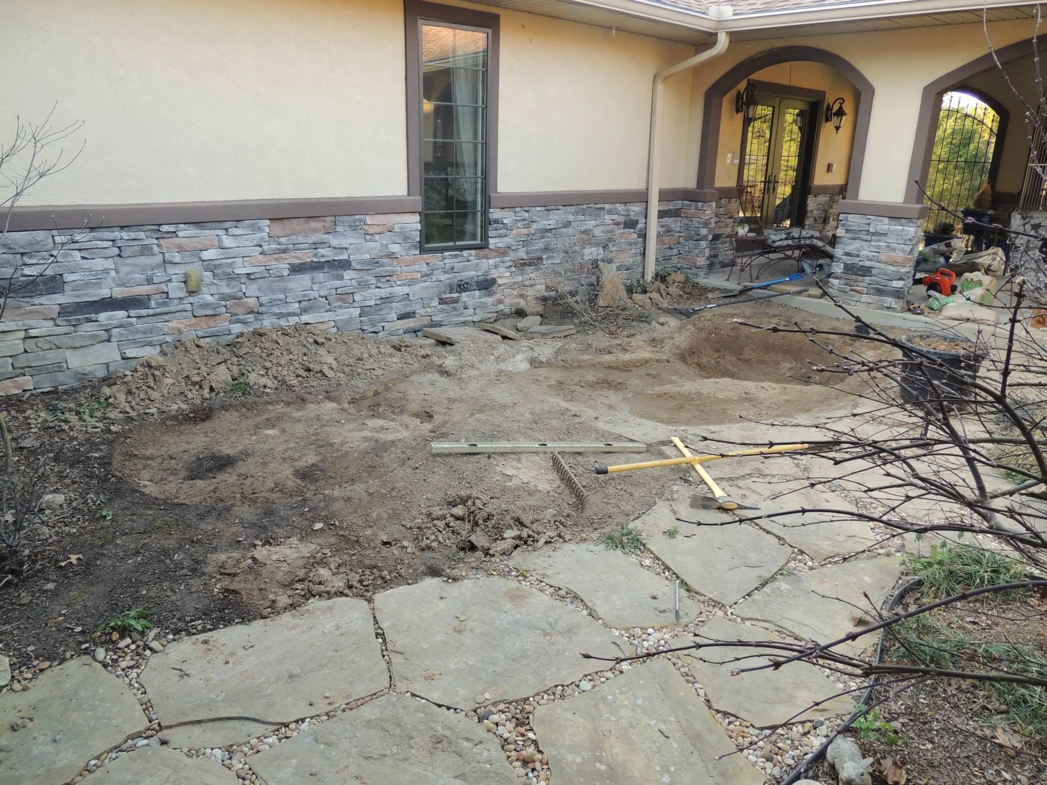 Front yard under construction with exposed soil, a concrete sidewalk, and a stone accent on the house wall. There are gardening tools, a bench, and construction materials near the porch.