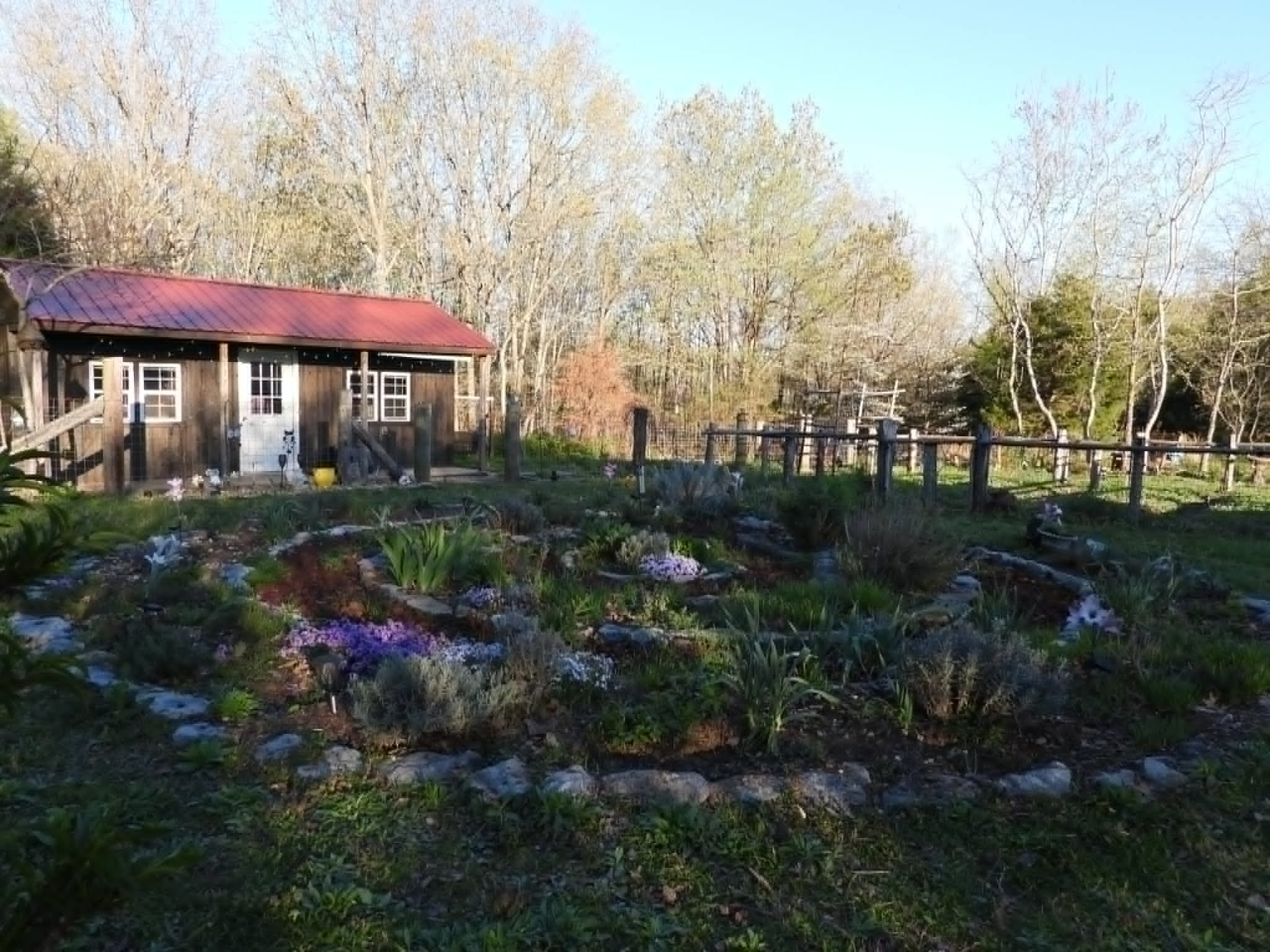 A rustic wooden shed with a red metal roof surrounded by a garden with flowers and plants, fenced in with a wooden fence and trees in the background under a clear blue sky.