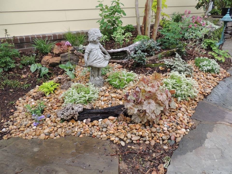 A small garden with various plants, a stone figurine of a girl, wooden logs, and a bright blue garden stake, surrounded by a border of small smooth stones and a stone pathway.