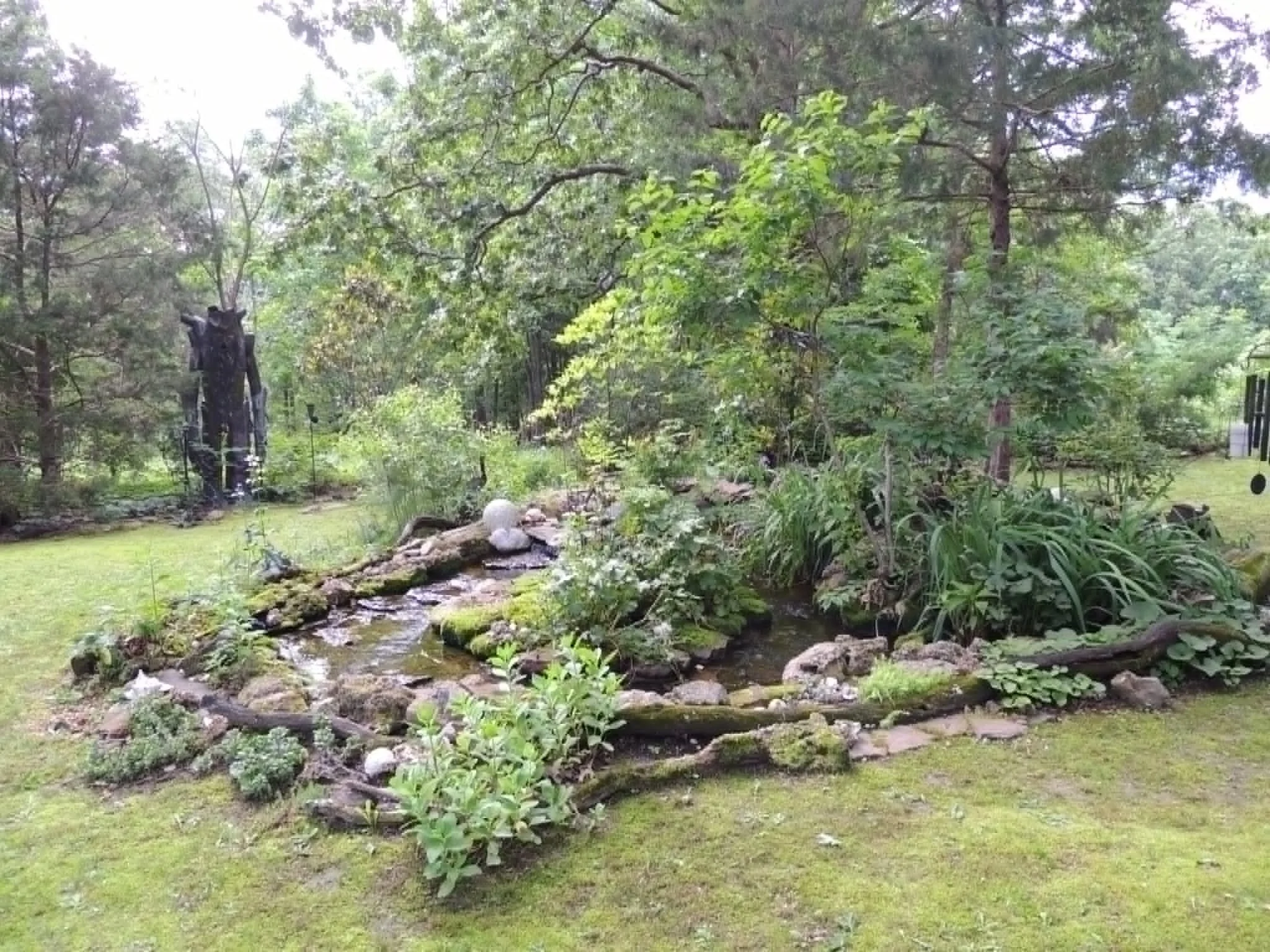 A landscaped backyard garden with a small creek, stone border, lush greenery, trees, and a lawn area.
