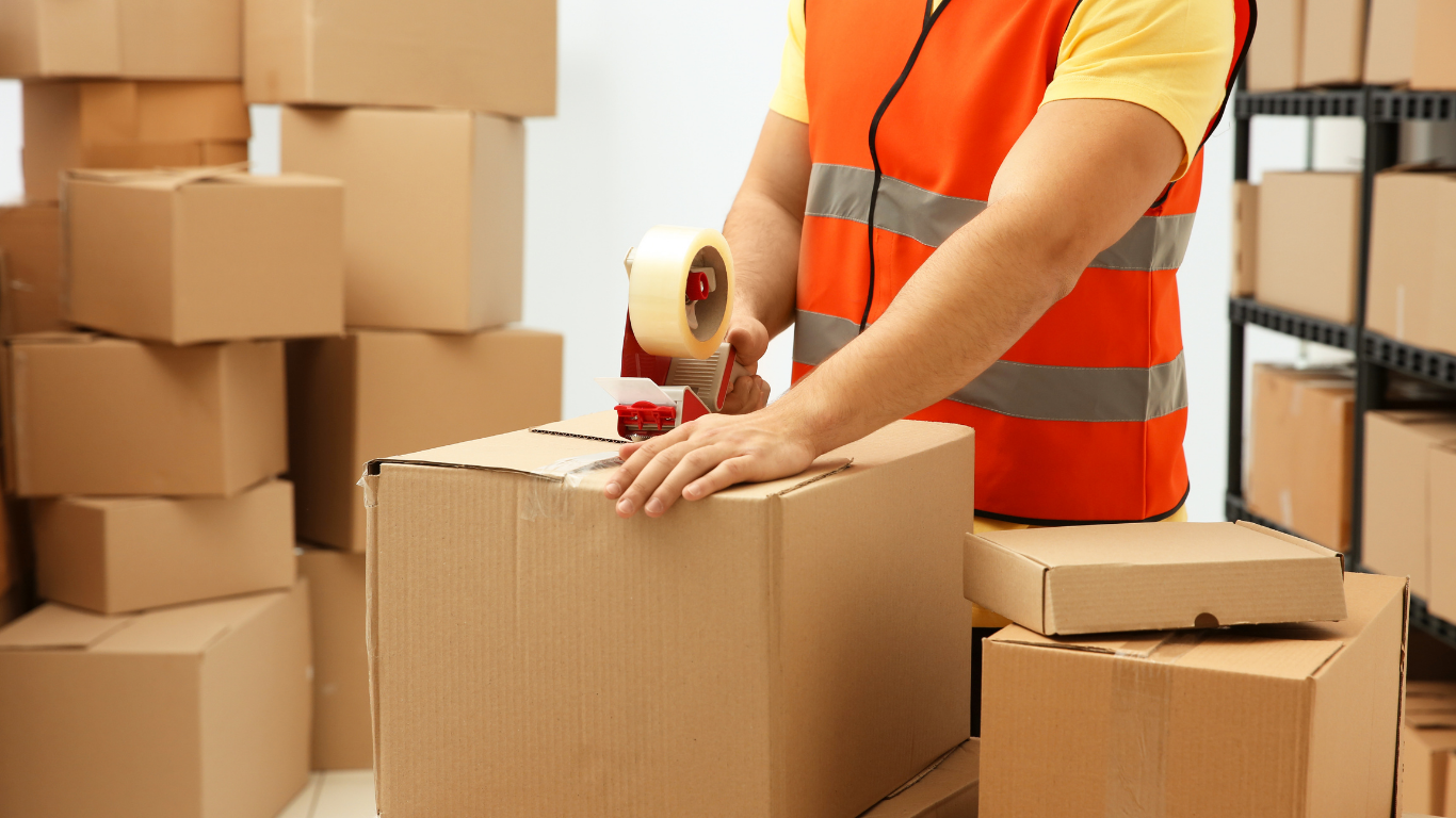 Person in a safety vest sealing a cardboard box with packing tape in a warehouse with shelves of boxes.