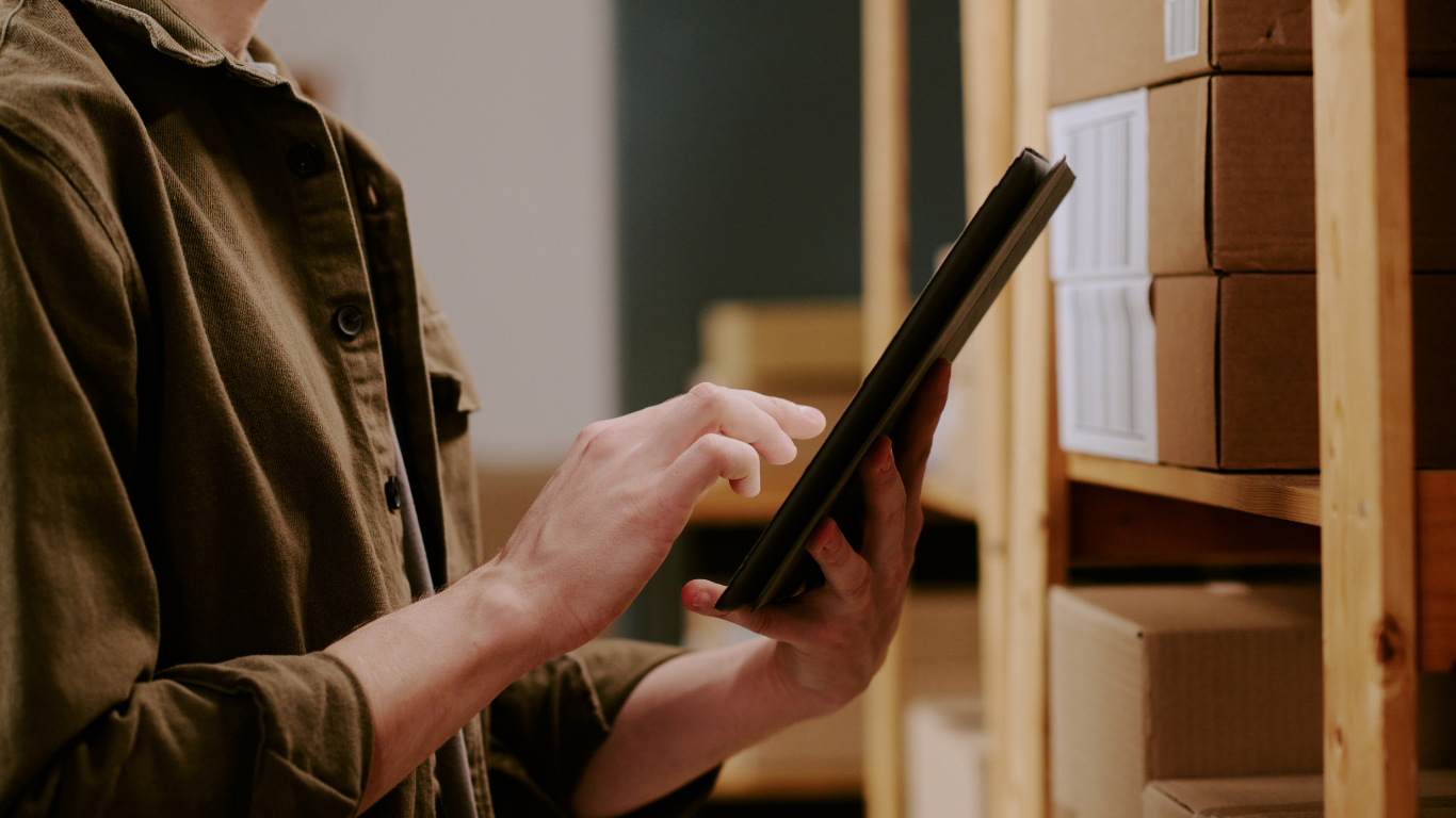 Person in brown jacket using a tablet in front of shelves with boxes.