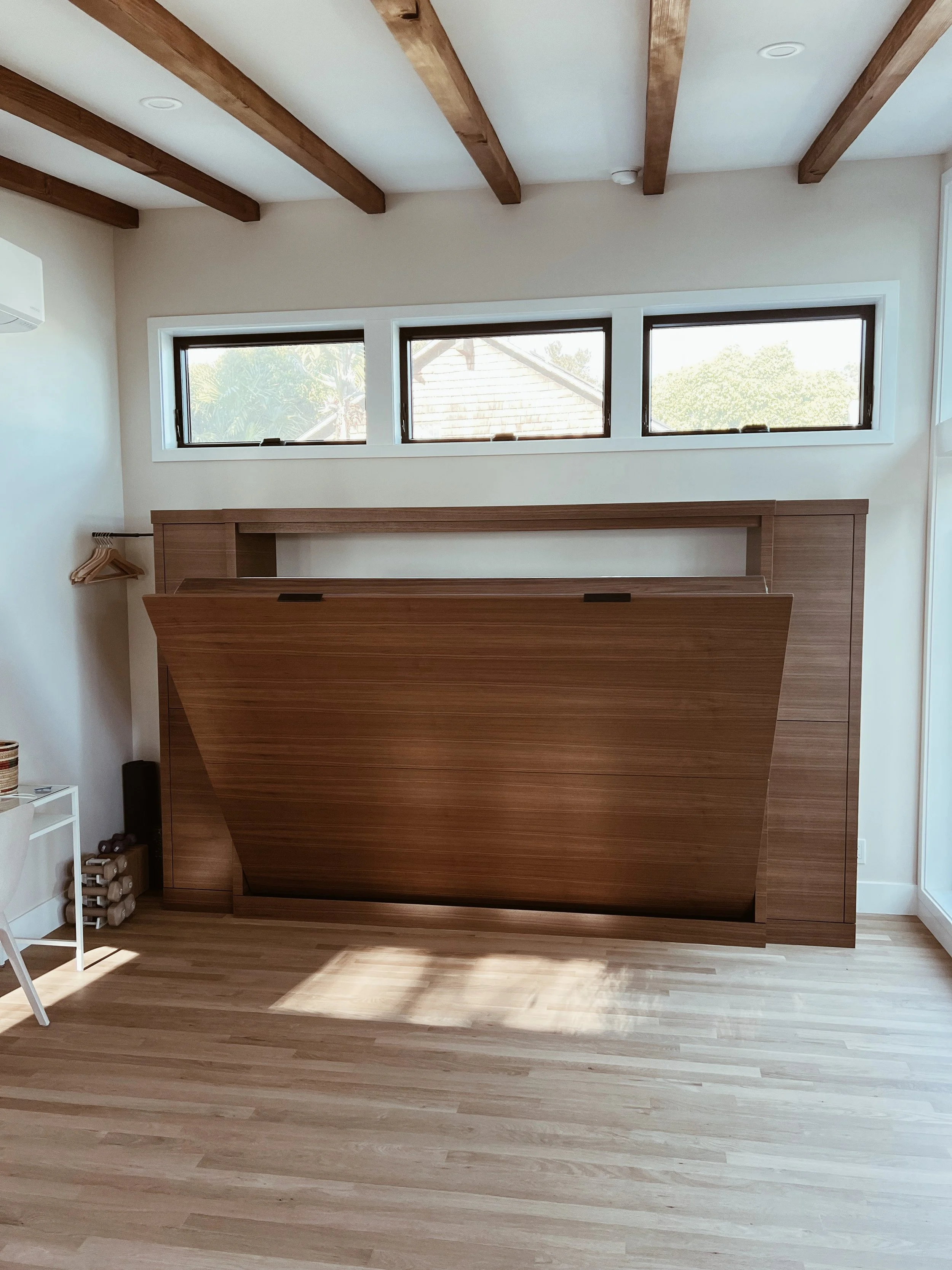 A room with three rectangular horizontal windows, a wooden floor, and a wooden piece of furniture that is open at the top. There is a white shelf with bottles and a small black speaker on the side, and a wine rack with bottles beneath it. The ceiling