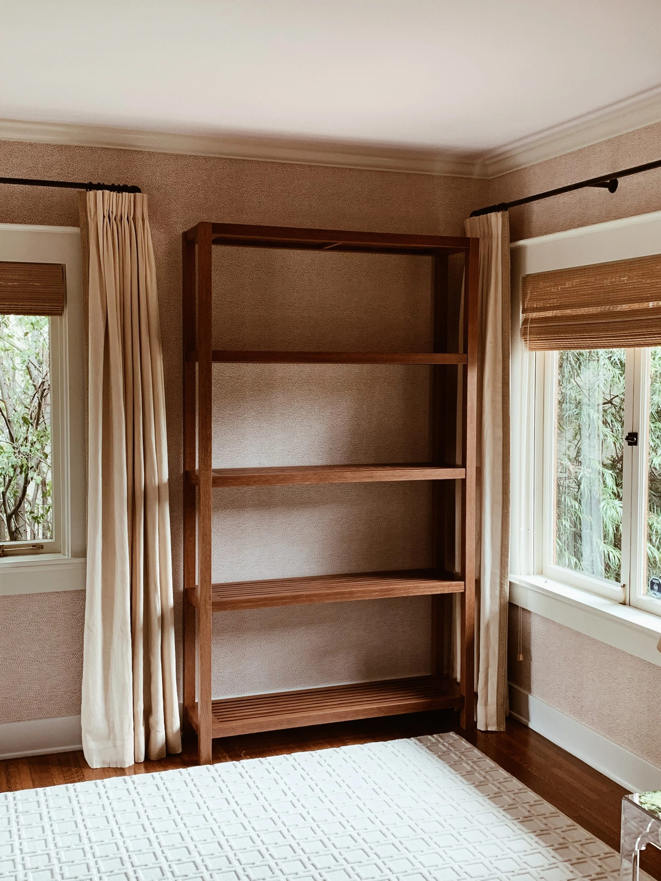 A wooden bookshelf standing next to a window with cream curtains and brown blinds in a room with textured beige walls and dark hardwood floors. Part of a white patterned rug is visible on the floor.