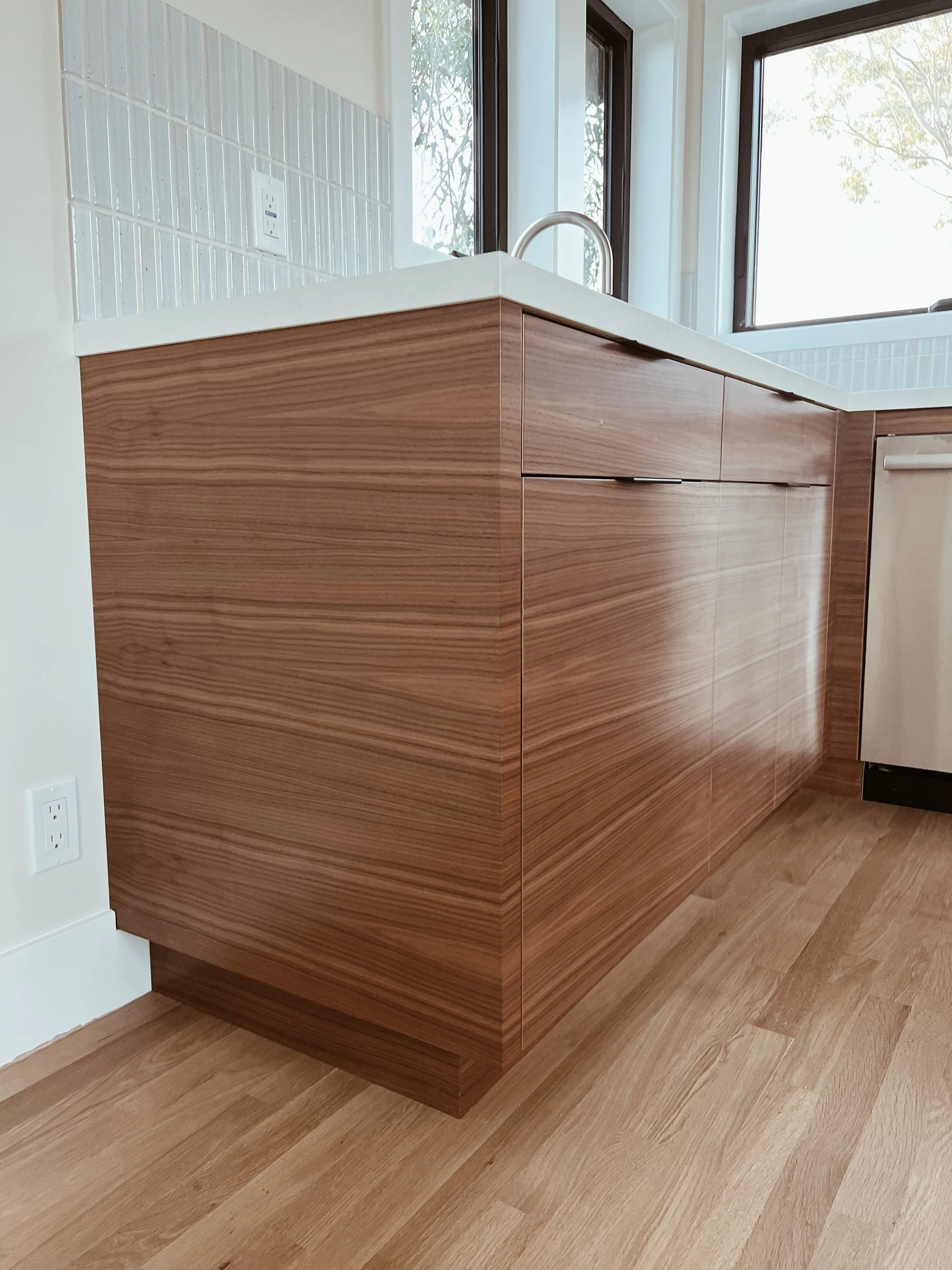 A modern kitchen corner with wooden cabinets, a white countertop, a stainless steel sink, and a black framed window with natural light coming in, showing trees outside.