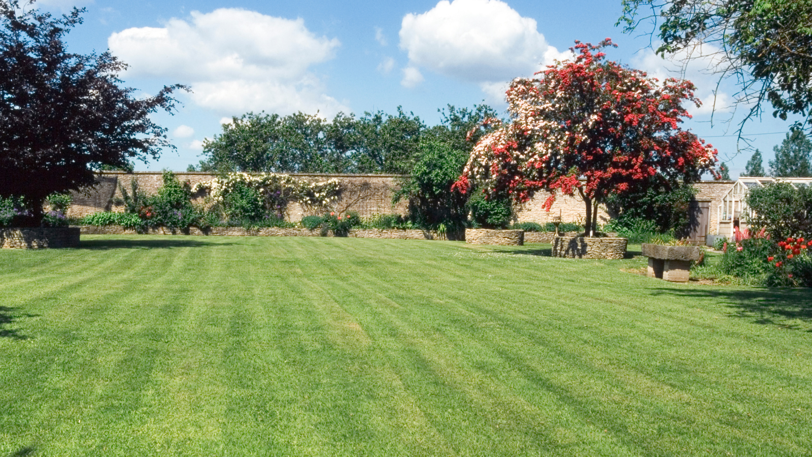 A lush green lawn with mowed grass, bordered by trees and flowerbeds, under a partly cloudy blue sky.