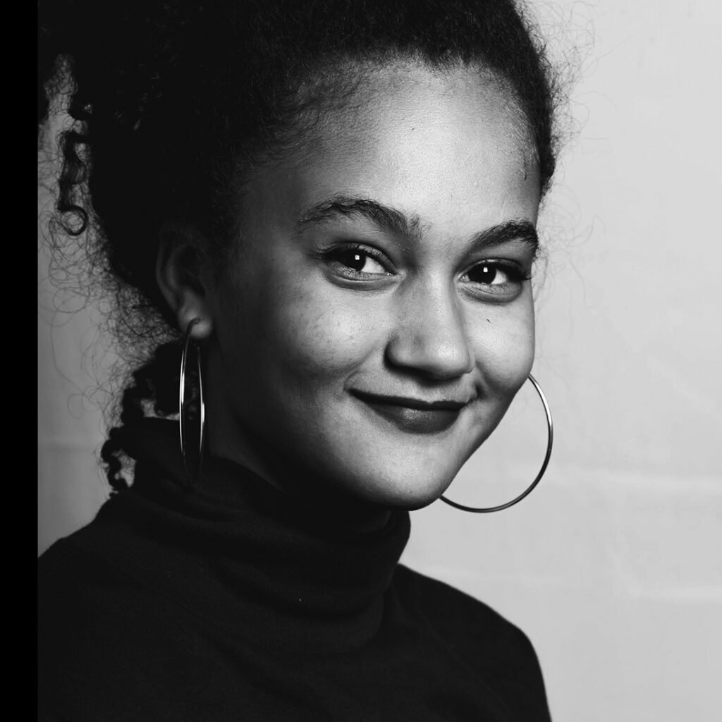 Black and white portrait of a woman with curly hair, hoop earrings, and a confident smile, wearing a turtleneck top.