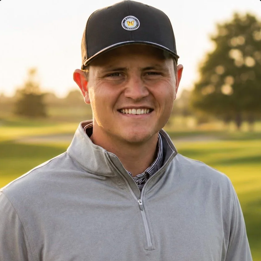 A young man smiling outdoors on a golf course during sunset, wearing a gray zip-up jacket and a black cap with a logo.