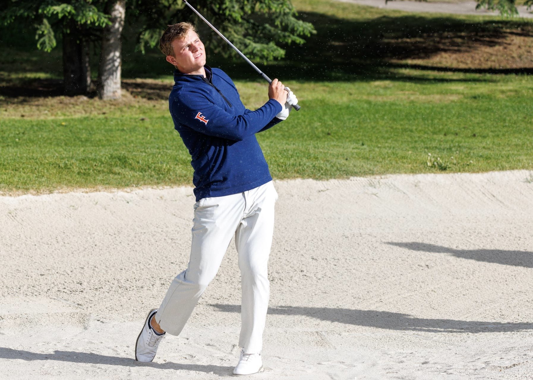 Young man in a blue jacket and white pants swinging a golf club on a golf course with sand trap, green grass, and trees in background.