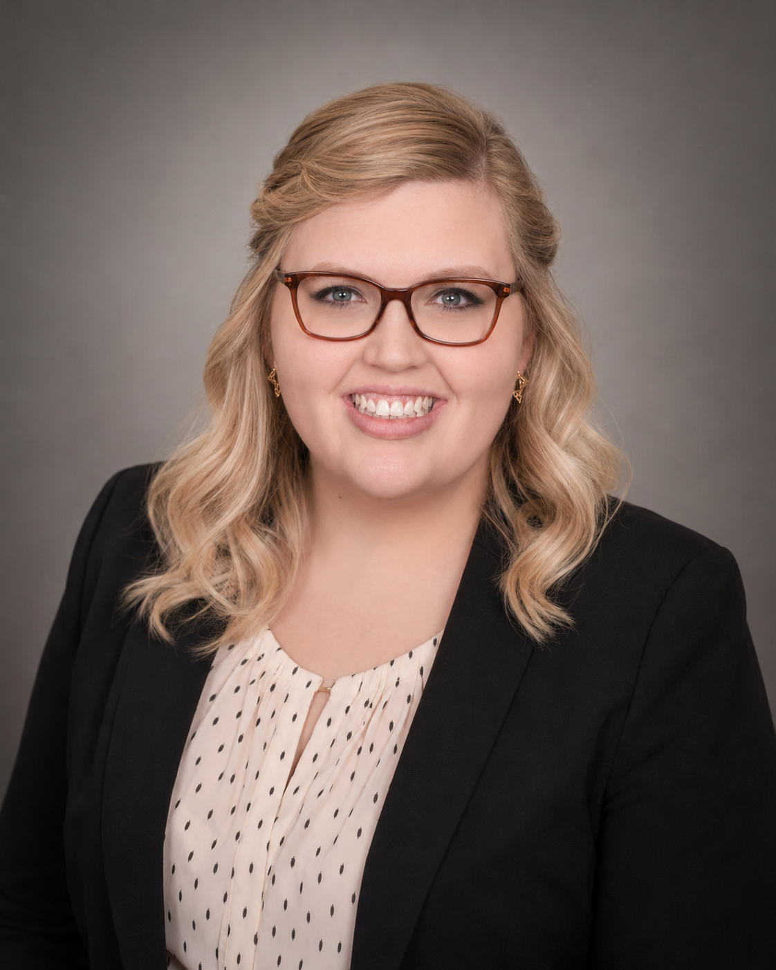 Professional headshot of a woman with blonde wavy hair, wearing glasses, gold earrings, a black blazer, and a white blouse with black polka dots against a gray background.